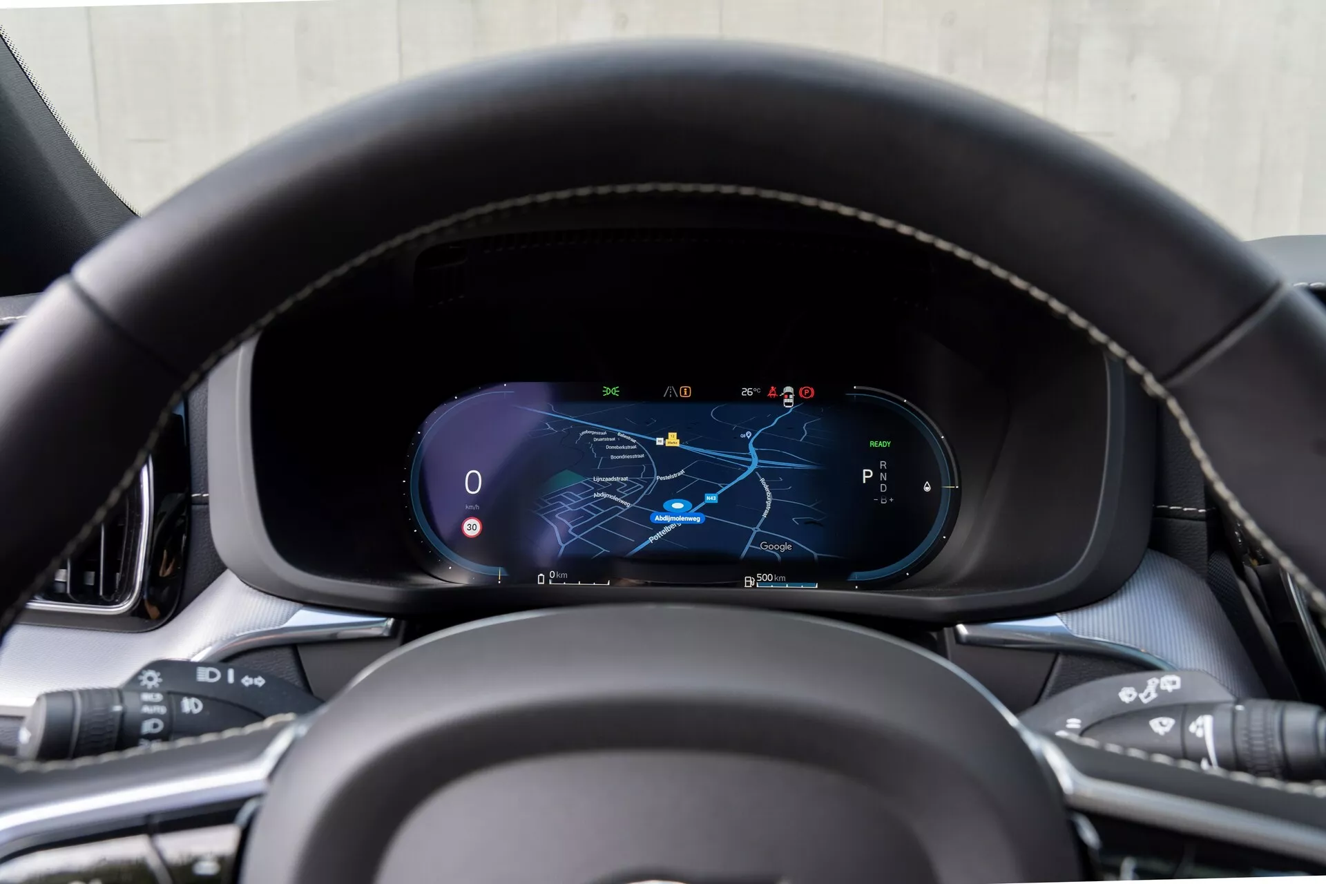 Vista ravvicinata del cruscotto della Volvo XC60 2021 (2a generazione restyling) dalla prospettiva del sedile del conducente, focalizzata sul quadro strumenti digitale con volante in pelle cucito a mano e pulsanti multifunzione per media, comandi vocali, controllo della velocità di crociera e sistemi di assistenza alla guida. Il display digitale mostra una mappa con il percorso di navigazione evidenziato, tachimetro, contagiri e selezione delle marce in modalità parcheggio, circondato da materiali interni di alta qualità.