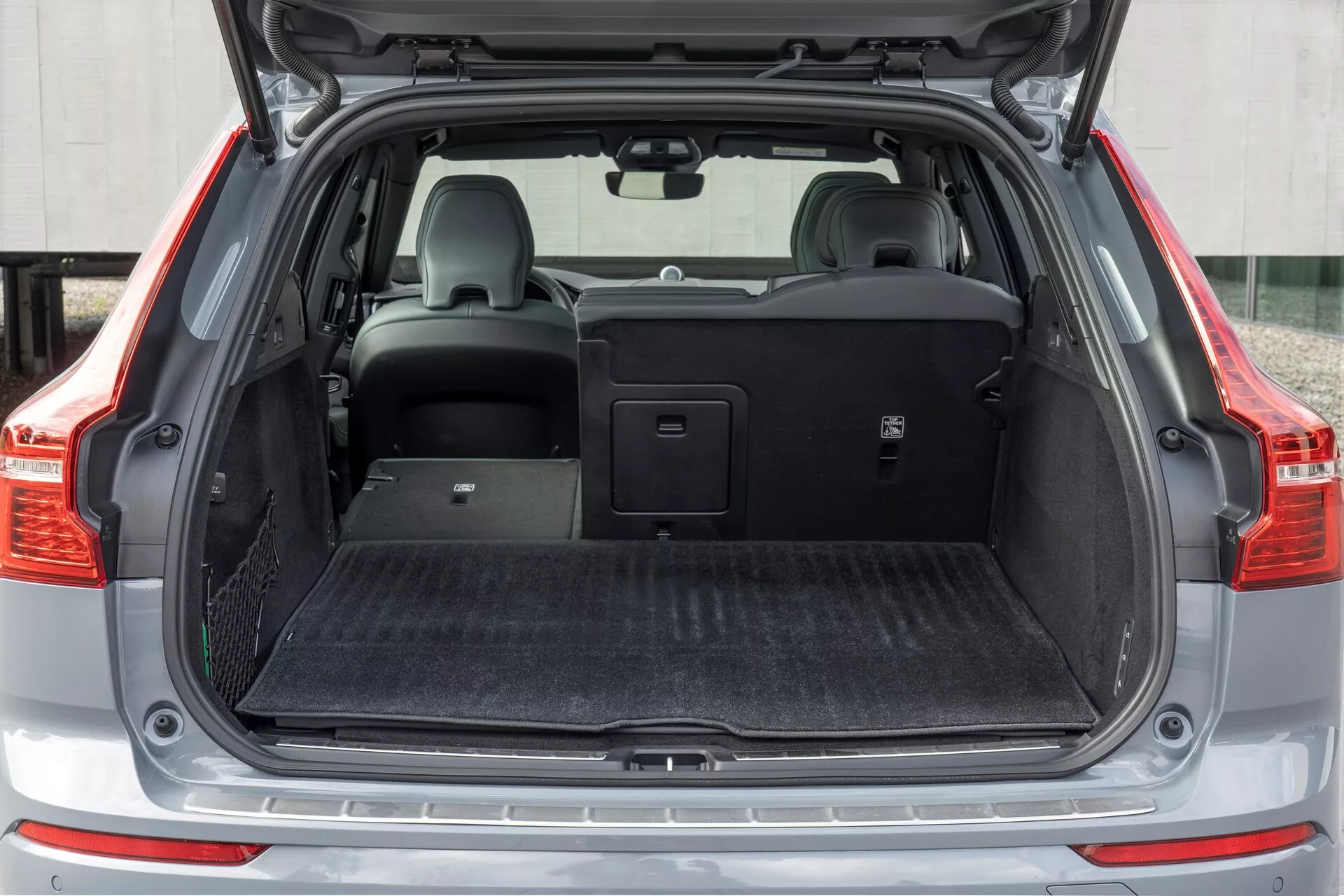 L'immagine mostra l'area di carico posteriore della Volvo XC60 2021 (facelift della seconda generazione), vista da un'angolazione frontale con il bagagliaio aperto. La ripresa a media distanza cattura l'intera larghezza dello spazio di carico, con un piano di carico piatto coperto da un materiale simile a moquette nera. Sul lato sinistro c'è un piccolo scomparto con una rete. Il sedile posteriore destro è abbassato, rivelando la capacità di piegatura divisa, mentre il sedile sinistro rimane in posizione verticale con un poggiatesta. Una copertura retrattile per il carico è posizionata sopra i sedili posteriori, illustrando la versatilità di stivaggio del veicolo.