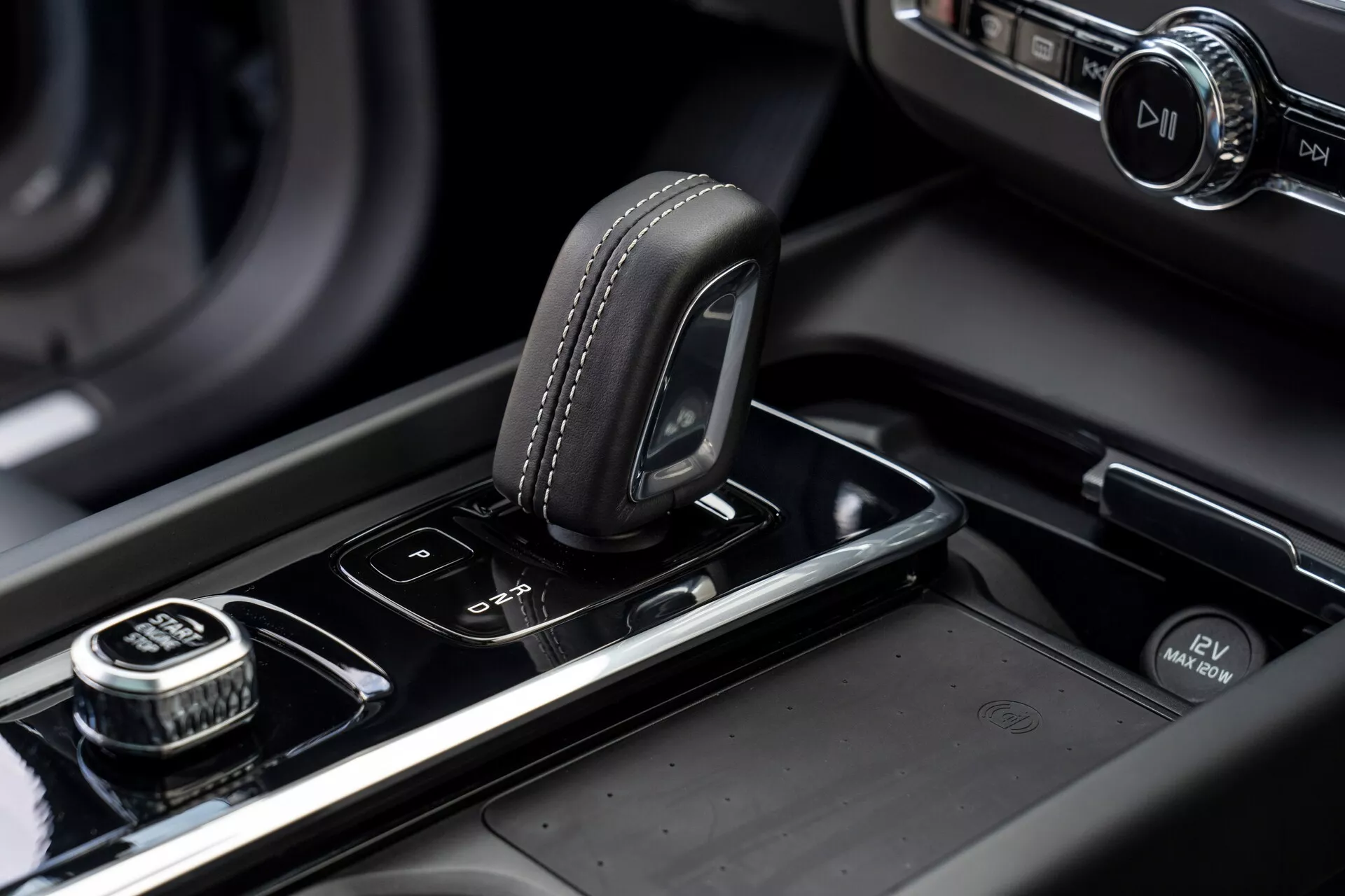 Primo piano della consolle centrale di una Volvo XC60 del 2021 (2a generazione, facelift). L'immagine evidenzia il selettore del cambio con parte superiore in pelle e dettagli di cucitura, situato su una superficie nera lucida. A destra c'è una manopola con bordo zigrinato, probabilmente per la navigazione del sistema di infotainment, con un pulsante di selezione a pressione. Accanto al selettore del cambio ci sono pulsanti per i sensori di parcheggio e il sistema start/stop automatico. La consolle centrale presenta rifiniture metalliche e uno schermo di infotainment illuminato sullo sfondo sfocato, il tutto mettendo in risalto il design moderno e di alta gamma del veicolo e la sua manifattura.