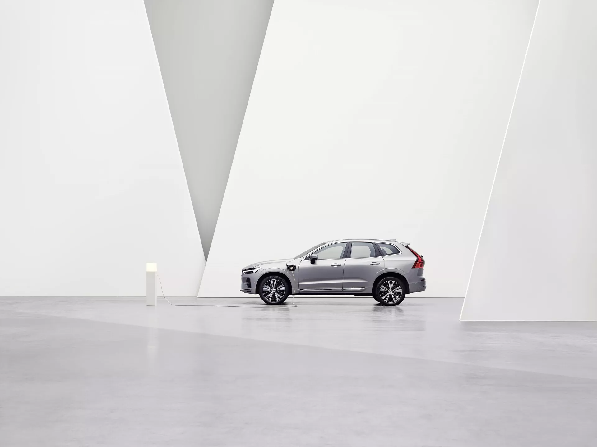 L'immagine presenta una Volvo XC60 2021 facelift di seconda generazione in una spaziosa e minimalista galleria d'arte con pareti bianche e pavimento lucido. Il veicolo, visto di profilo verso sinistra, mette in evidenza le ruote in lega, le linee caratterizzanti delle porte laterali, gli specchietti retrovisori, i vetri oscurati e le distintive barre sul tetto. Da un angolo leggermente basso sono visibili la griglia frontale, i fari a LED, le frecce integrate e i sensori di parcheggio del paraurti anteriore. L'ambiente pulito e la parete con partizione a forma di V sottolineano il design dell'auto e la sua posizione premium sul mercato sotto una luce soffusa e diffusa.