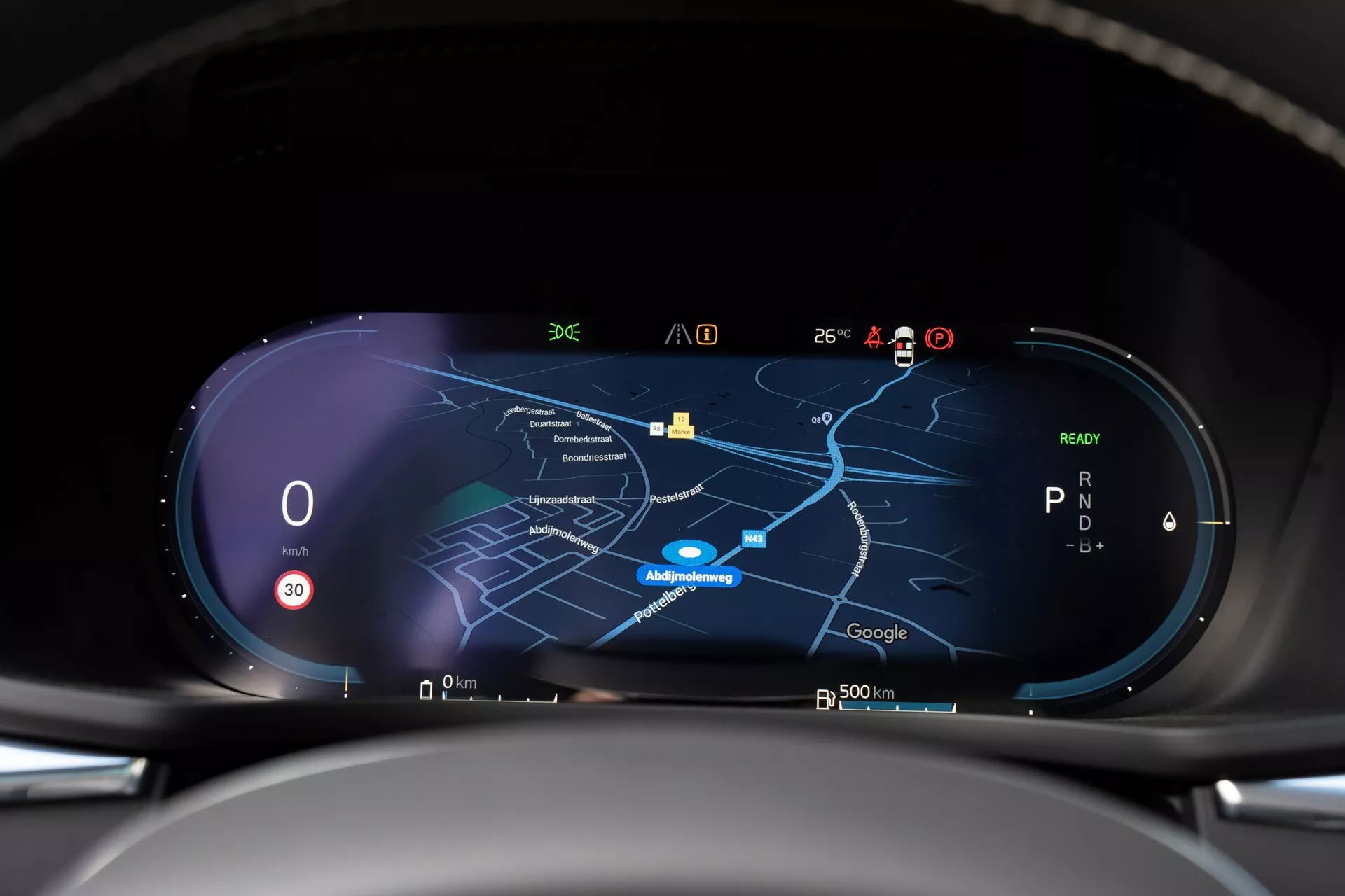 Vista interna della Volvo XC60 2021 di seconda generazione con restyling, che mostra il cruscotto digitale ad alta risoluzione dalla prospettiva del conducente. Il cruscotto presenta un display widescreen con tachimetro che segna zero, un indicatore di marcia che mostra 'P' e una mappa di navigazione centrale in modalità notturna. Vari indicatori per ricezione del segnale, temperatura esterna, carica della batteria e funzioni di assistenza alla guida sono visibili lungo il bordo superiore, indicando connettività in tempo reale e capacità tecnologiche avanzate.