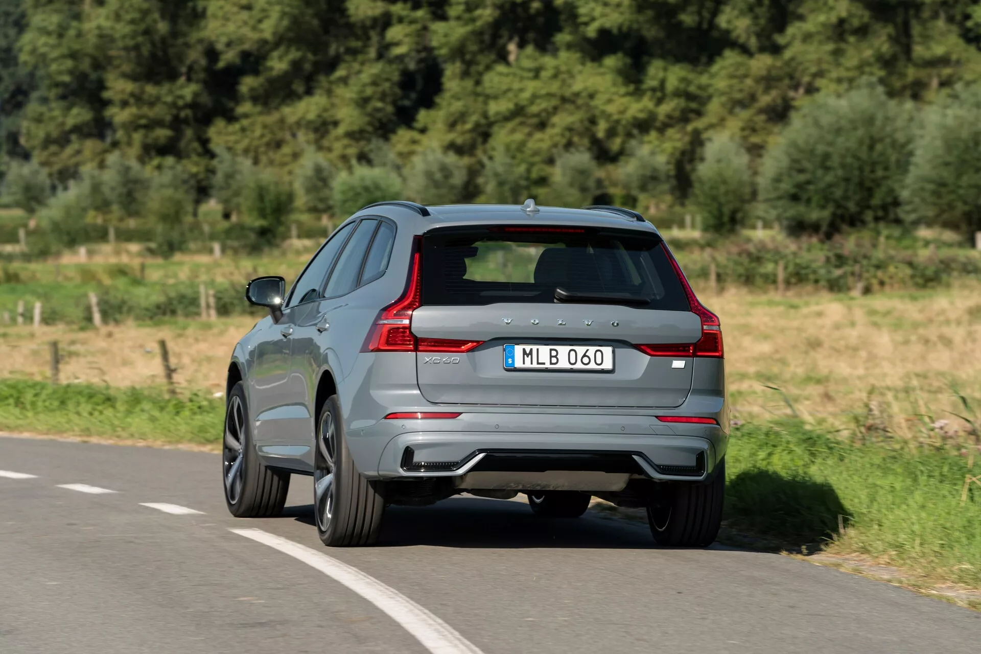 Vista tre quarti posteriore di una Volvo XC60 2021 che si muove lungo una strada asfaltata. Catturata da un'angolazione elevata, questo moderno SUV mostra la sua vernice grigia lucida, le distintive luci posteriori a LED, i doppi terminali di scarico e il diffusore posteriore. Il badge del produttore e la designazione del modello sono visibili sul portellone scolpito, completati da finestrini oscurati e cerchi in lega con design a più razze, suggerendo una versione più sportiva. L'immagine offre una visione chiara del design aerodinamico e delle caratteristiche di alto livello tipiche di un veicolo moderno.