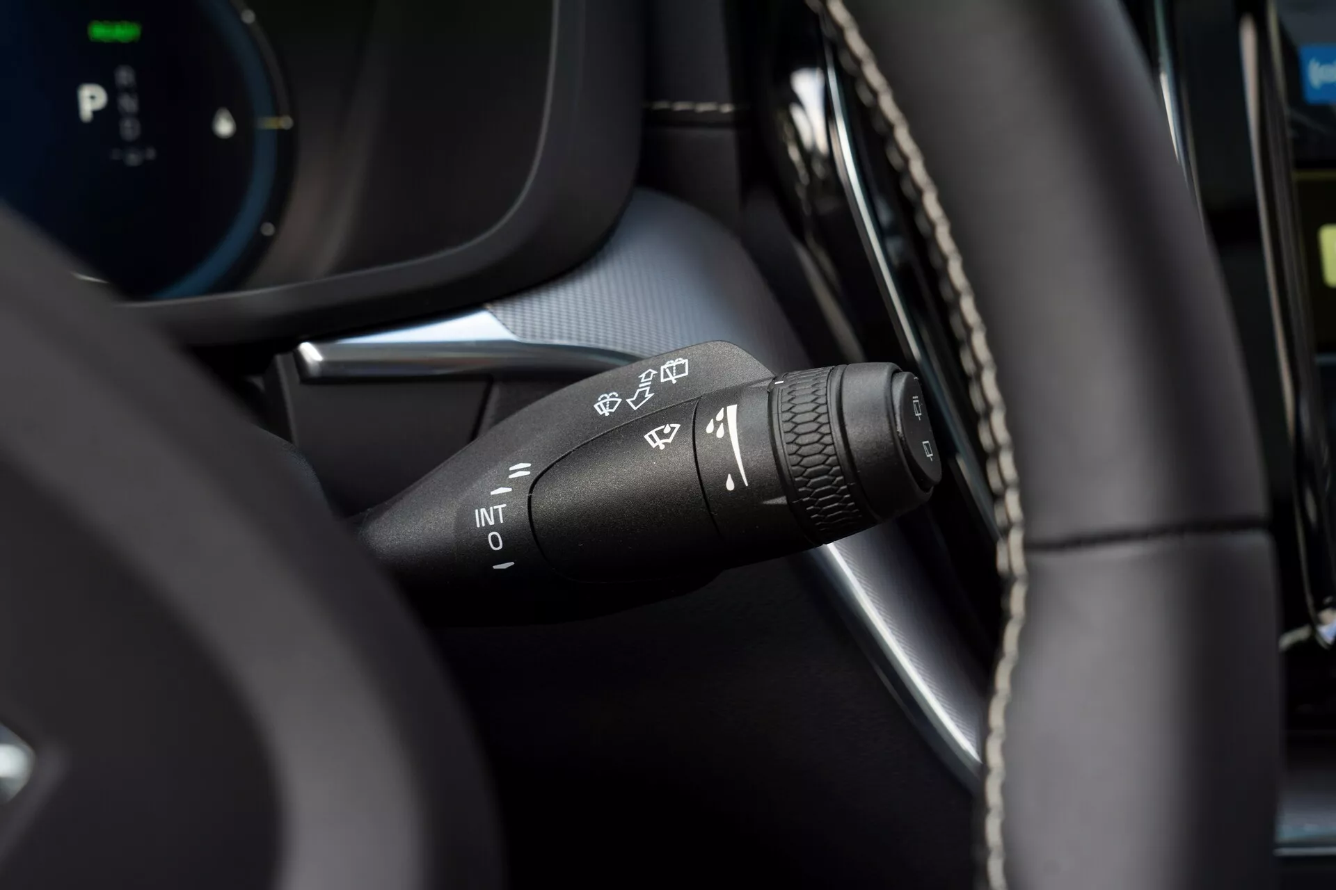 Primo piano della leva di controllo del tergicristallo della Volvo XC60 2021 (facelift della seconda generazione) dalla prospettiva del conducente. Messa a fuoco sui vari simboli delle impostazioni del tergicristallo: rilevamento automatico della pioggia, velocità intermittente, bassa e alta. La punta della leva presenta un anello rotante per regolare l'intervallo dei tergicristalli intermittenti. Interni moderni con una combinazione di materiale simile alla pelle e componenti in plastica nera, con un cruscotto digitale sfocato sullo sfondo.