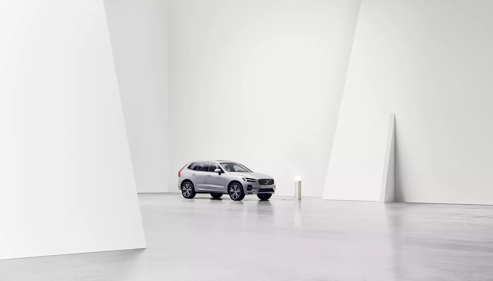 Immagine di una Volvo XC60 2021 (2ª generazione, facelift) posizionata centralmente in un ambiente da studio minimalista, spazioso e ben illuminato, ripresa da una prospettiva anteriore a tre quarti. Il colore argento chiaro dell'auto contrasta con le ruote più scure e una parte della griglia anteriore, mostrando elementi di design moderni di SUV come linee curve e angolari. La vista evidenzia la griglia anteriore, i fari, il cofano, il parabrezza e il lato del conducente, comprese le porte anteriori e posteriori e i finestrini laterali. Lo sfondo bianco nitido accentua l'auto, focalizzando l'attenzione sul suo design e le sue caratteristiche, adatto per scopi promozionali o artistici.