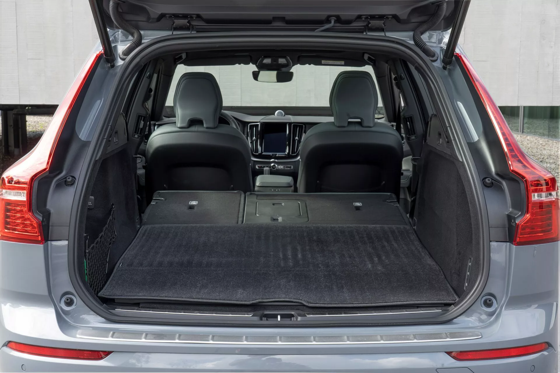 Area di carico posteriore di una Volvo XC60 2021 (seconda generazione facelift) da una prospettiva posteriore diretta. Lo spazio aperto del bagagliaio rivela un pavimento di carico piatto, rivestito in moquette e vuoto, con i sedili posteriori abbattuti per aumentare la capacità di stivaggio. L'interno è moderno, con un tappetino protettivo sul pavimento, ma senza strumenti organizzativi visibili come reti da carico o ganci.