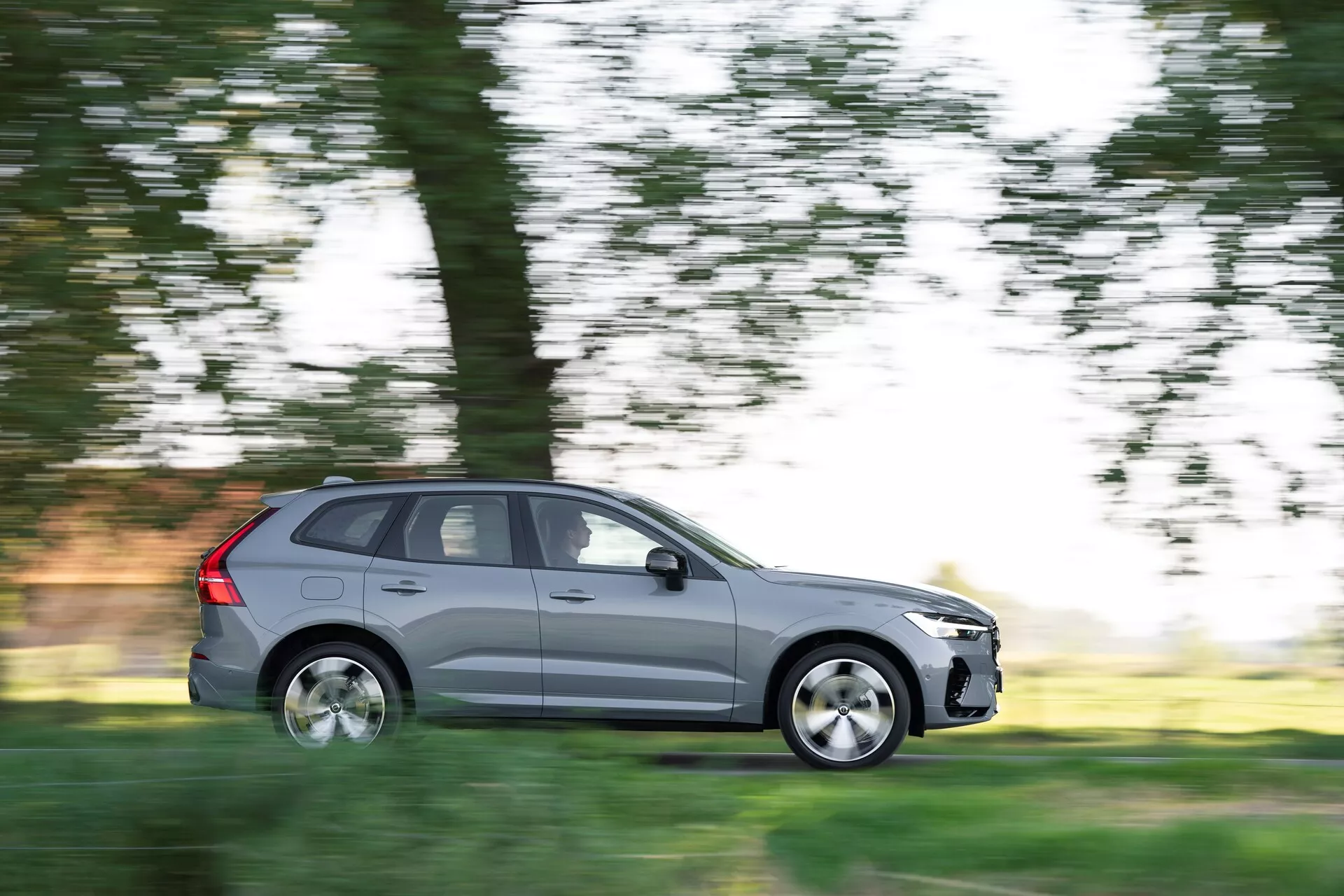 L'immagine mostra un SUV Volvo XC60 2021 (2a generazione, facelift) in movimento, catturato con una tecnica di panning che sfoca lo sfondo mantenendo l'auto a fuoco. Vista dal lato del conducente in un profilo laterale completo, il veicolo grigio metallizzato presenta cinque porte, cerchi in lega e pneumatici standard per la strada. La scena suggerisce una guida diurna in un ambiente naturale, con alberi ed erba sfocati sullo sfondo, enfatizzando il movimento dell'auto.