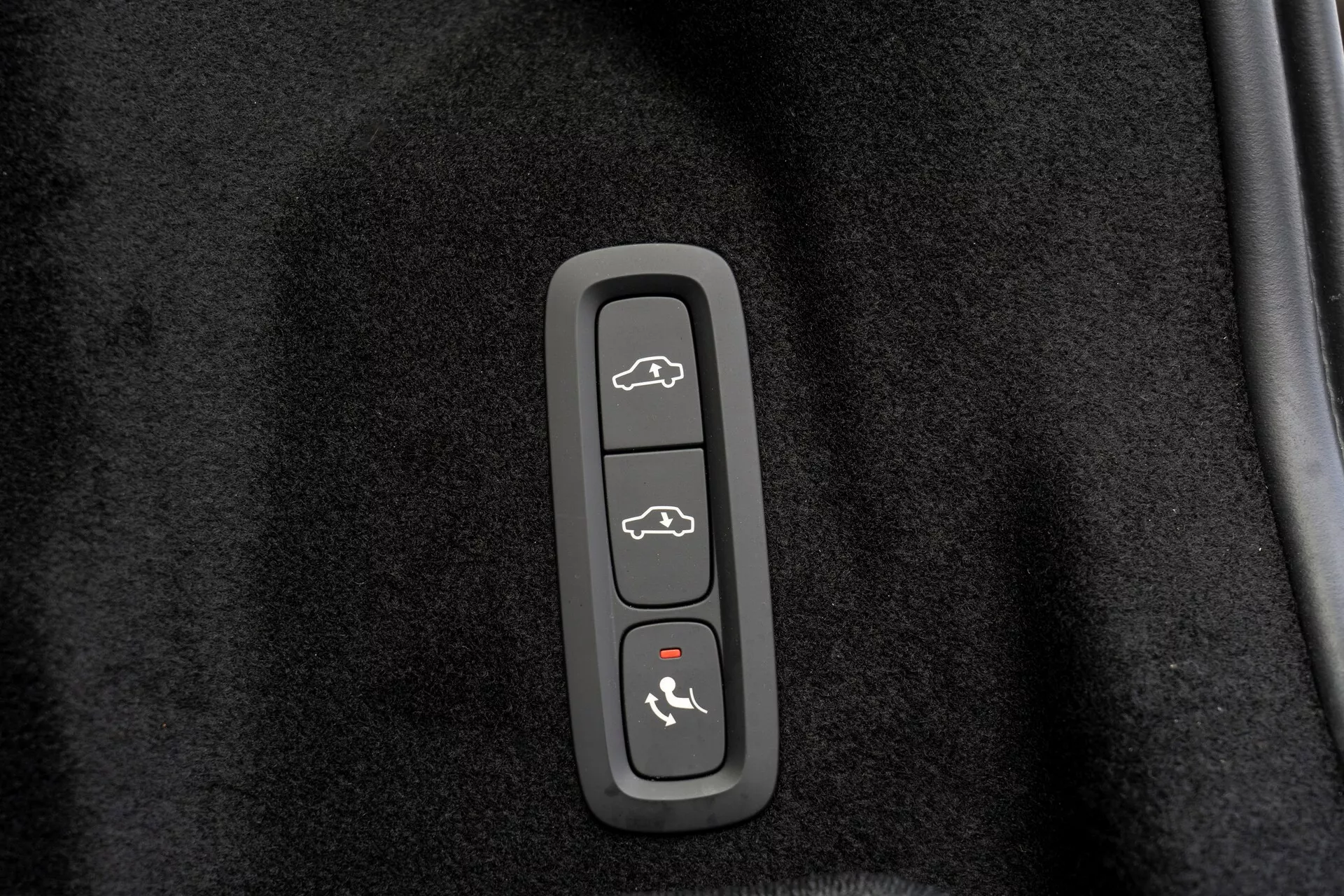 Primo piano di un pannello di controllo in una Volvo XC60 2021 (2ª generazione, facelift), incorporato in una modanatura di plastica su uno sfondo nero simile a un tappeto. Ha quattro pulsanti: in alto - veicolo con il bagagliaio aperto, secondo - siluetta dell'auto con tutte le porte aperte, terzo - auto con una freccia che punta verso il cofano, in basso - auto con una linea curva sopra, probabilmente indicando apertura del bagagliaio, blocco/sblocco delle porte, accesso al cofano/modalità valet e attivazione del sistema di sicurezza o allarme panico.