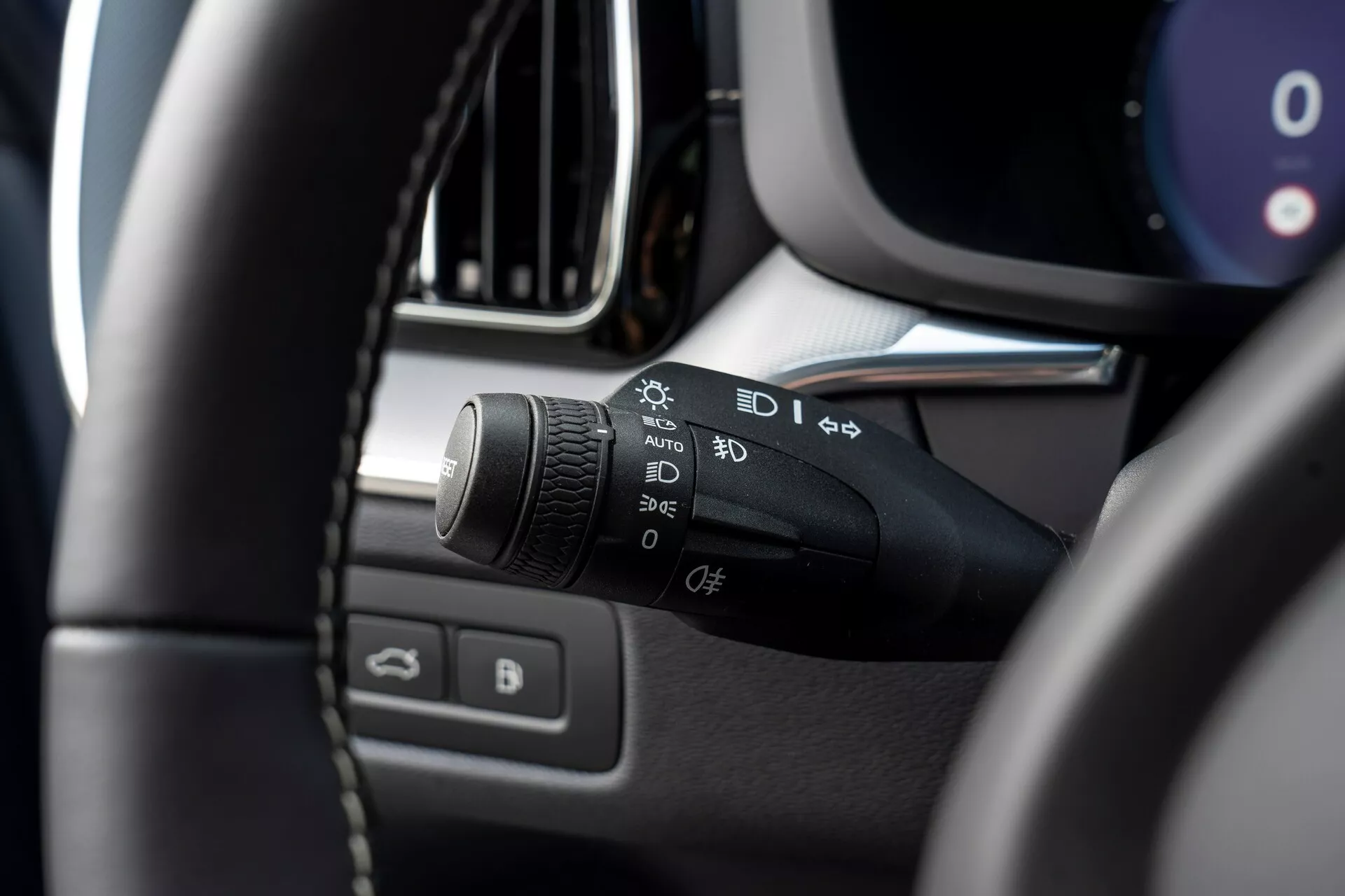 Inquadratura ravvicinata dell'interno di una Volvo XC60 2021 (seconda generazione facelift), focalizzata sulla colonna di sterzo laterale sinistra. Il soggetto principale è la leva di controllo dei fari e degli indicatori, con simboli per luci automatiche, luci di parcheggio, fari, impostazioni dei fendinebbia e una funzione personalizzabile. L'immagine è scattata dal sedile del conducente, leggermente verso l'alto e a sinistra, mostrando la parte in alto a sinistra del volante sfocata in primo piano e mettendo in risalto la leva di controllo. Altri dettagli sfocati includono parti del pannello strumenti, console centrale, bordo della ventilazione o schermo di infotainment e porta del conducente con comandi per finestre e specchietti.