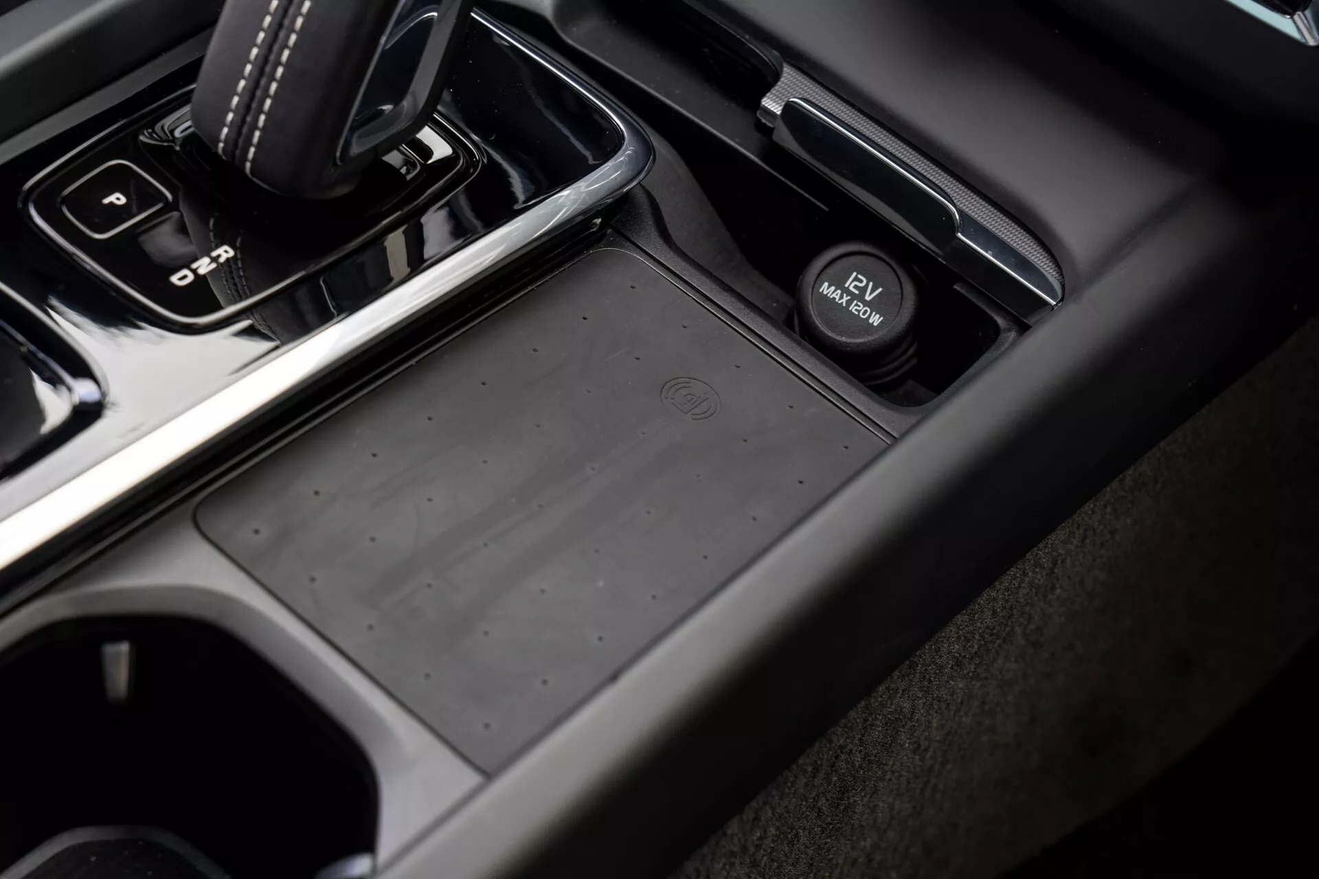 Vista ravvicinata e angolata dell'area della console centrale di una Volvo XC60 2021 (seconda generazione restyling) dal lato passeggero, con un cambio automatico in posizione 'park', un vano portaoggetti antiscivolo, un interruttore del freno di stazionamento elettronico, portabicchieri e una combinazione di materiali in plastica morbida al tatto e finitura metallica in tonalità scure e nere.