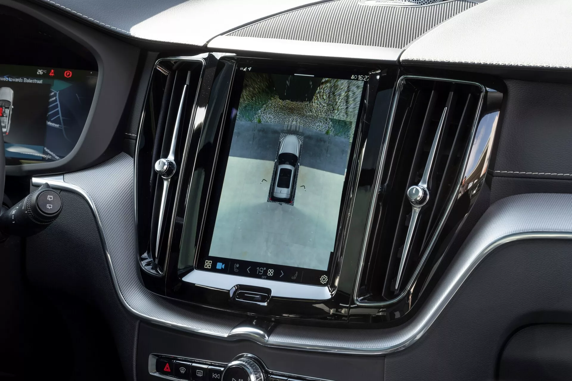 Vista interna di una Volvo XC60 2021 (restyling di 2a generazione) che mostra un grande schermo touch verticale di infotainment sulla consolle centrale, visualizzando il feed della telecamera posteriore con una vista dall'alto. Finiture di lusso con accenti cromati ed elementi di design elegante, comandi aggiuntivi sotto lo schermo e una vista parziale del volante, indicando un interno premium e tecnologicamente avanzato.