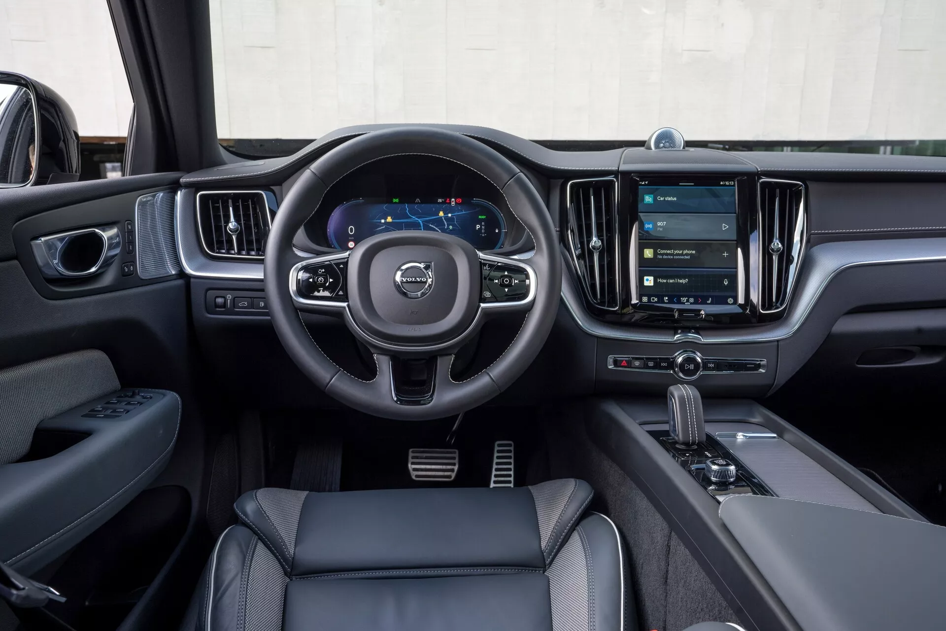 Interni di una Volvo XC60 2021 (restyling 2a generazione) dalla prospettiva del conducente. L'immagine mette in risalto il volante a tre razze con pulsanti di controllo per audio, comunicazione e impostazioni del cruise control, il cruscotto digitale che visualizza tachimetro e navigazione, e il grande sistema di infotainment a schermo verticale. Gli elementi di design moderno includono materiali simili alla pelle, accenti cromati e una leva del cambio automatico con selettore della modalità di guida. La porta del conducente mostra i comandi per finestrini e specchietti, che enfatizzano le caratteristiche tecnologiche e di lusso.