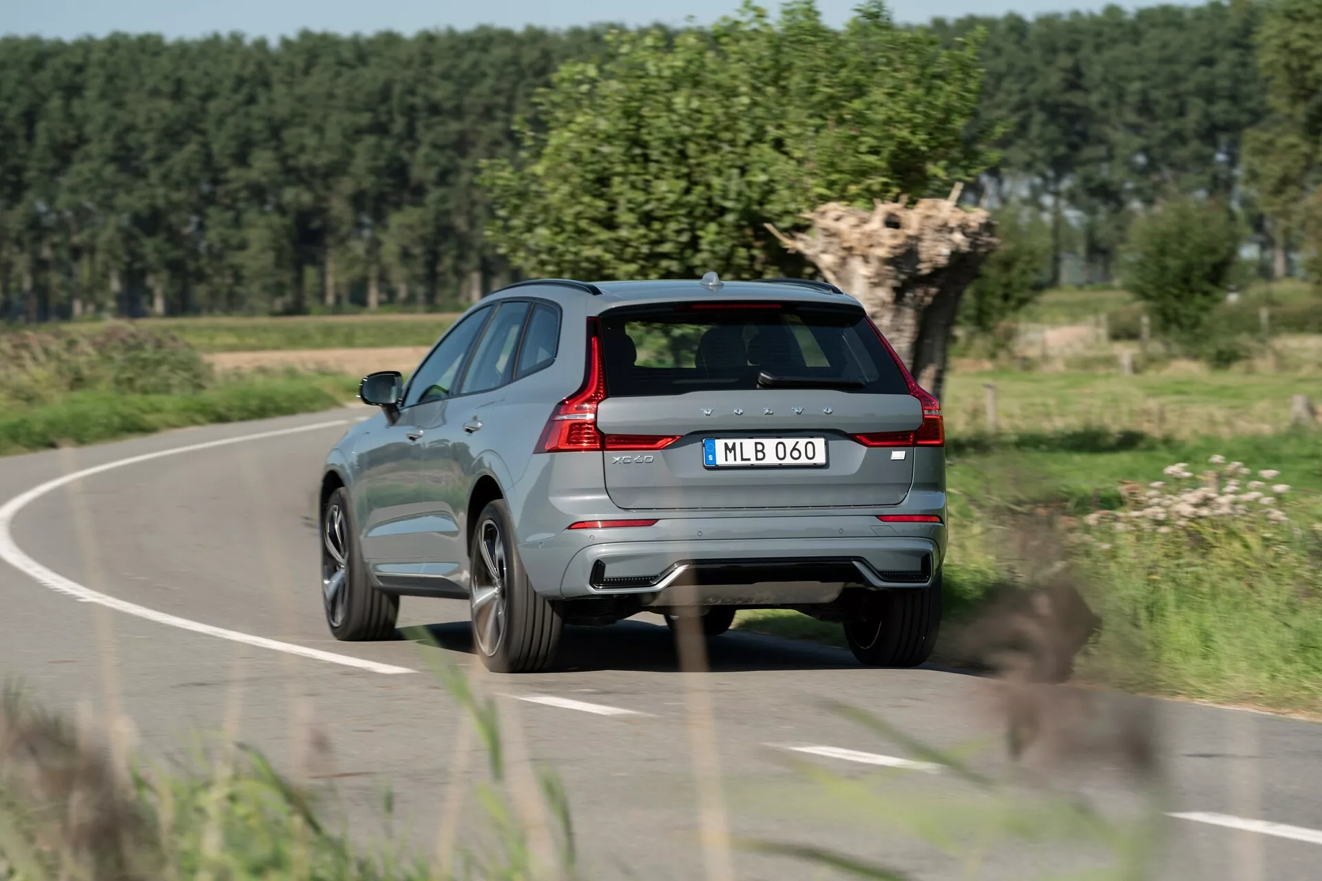 Vista posteriore tre quarti di un SUV Volvo XC60 2021 grigio metallizzato moderno in movimento su una strada di campagna, che mostra fanali posteriori distintivi, sistema di scarico doppio, tergilunotto posteriore, prominente spoiler posteriore, sensori integrati sul paraurti posteriore, targa centrale, cerchi in lega, linea di carattere affilata lungo il lato, finiture riflettenti delle finestre e sistema di guide sul tetto. Sfondi con fogliame sfocato e cielo sereno che indicano movimento e ambiente rurale.