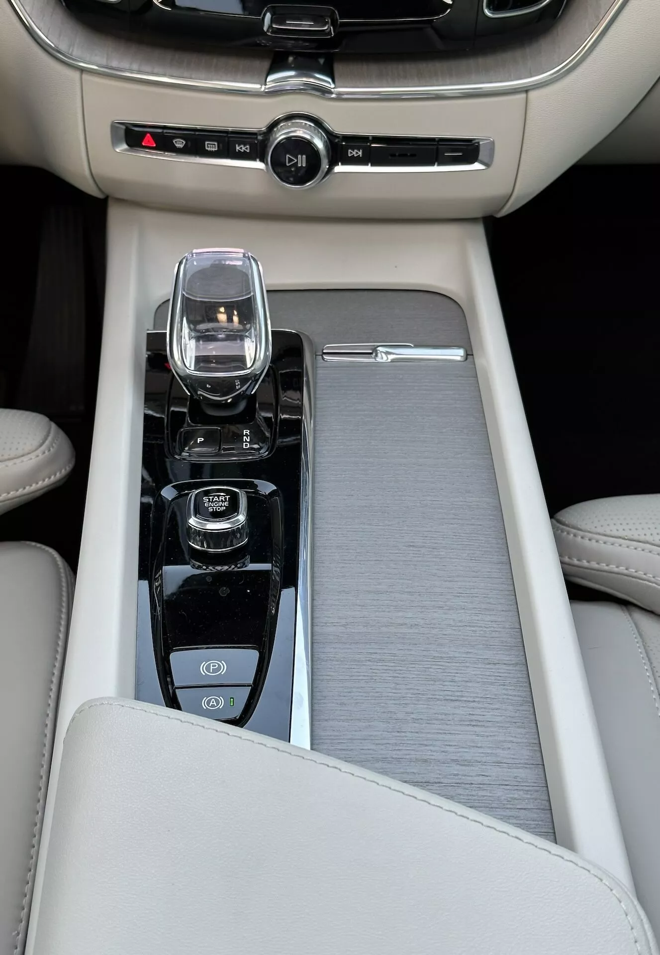 Primo piano della consolle centrale in una Volvo XC60 Recharge ibrida plug-in del 2021, con bracciolo in pelle beige, una finitura in metallo spazzolato scorrevole, un moderno controller per il sistema infotainment, un selettore di marcia in vetro cristallo per il cambio automatico, e comandi per avvio/stop motore, disabilitazione della trazione e selezione della modalità di dinamica di guida, insieme al controllo climatico bizona.