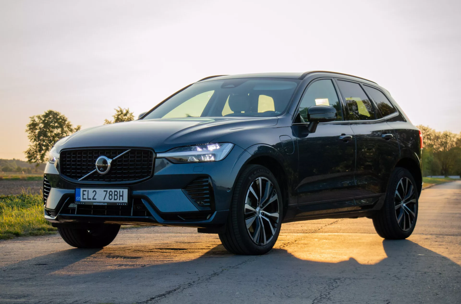 SUV moderno nero Volvo XC60 Recharge Plug-in Hybrid 2021 parcheggiato su una strada asfaltata all'alba o al tramonto, mostrando il suo design con focus sulla griglia, fari LED, ruote in lega e profilo laterale su sfondo di cielo sfumato