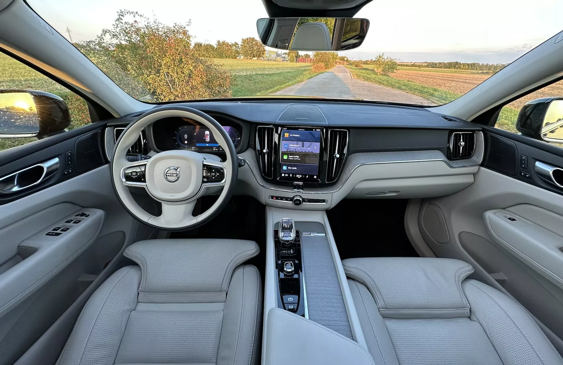 Vista interna di una Volvo XC60 Recharge Plug-In Hybrid 2021, presa dall'area dei sedili posteriori mostrando l'intero cruscotto, volante multifunzione con logo, pannello strumenti digitale, sistema di infotainment centrale touchscreen e sedili di lusso in pelle di colore chiaro.