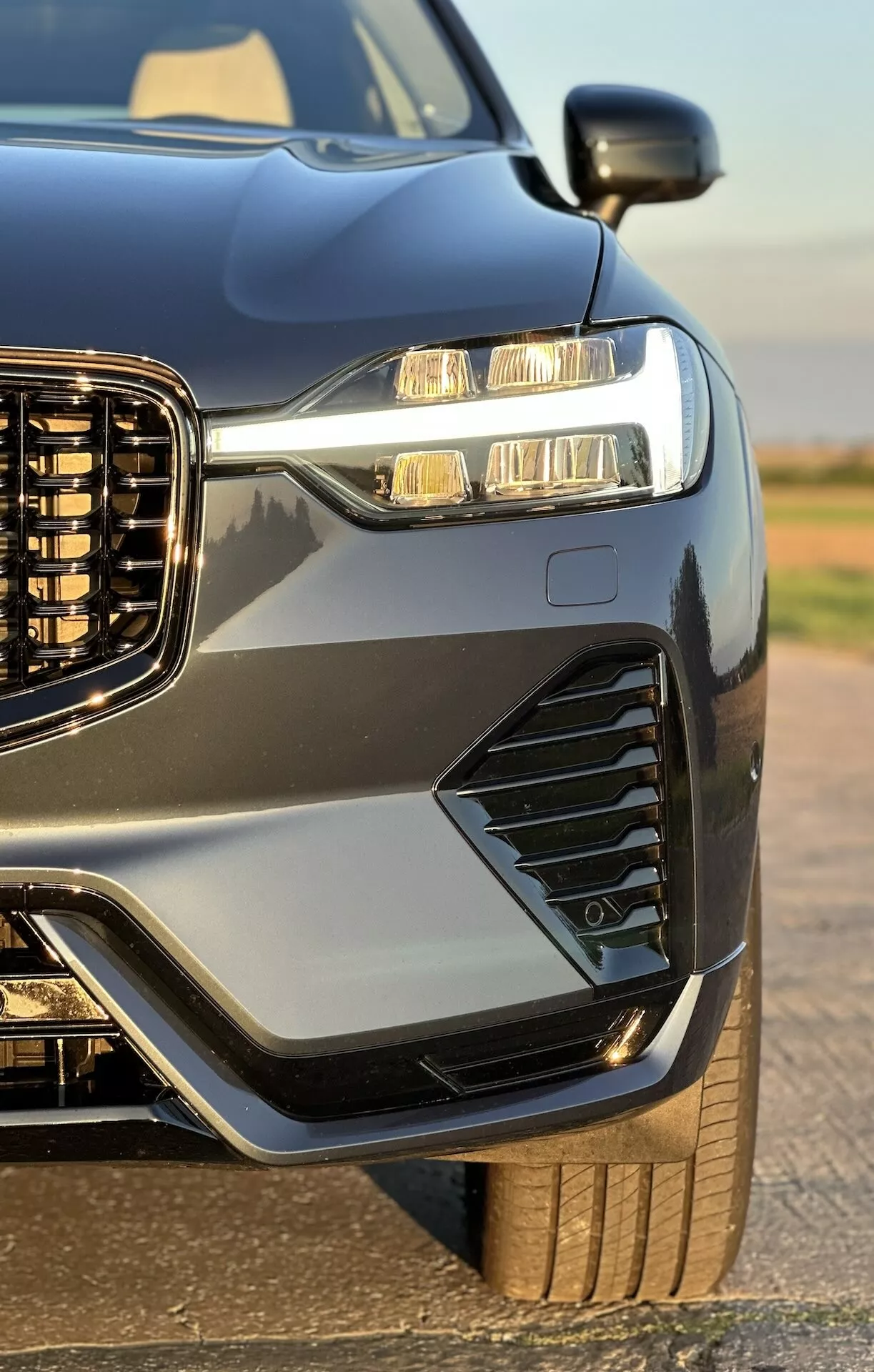 Vista ravvicinata del quarto anteriore di una Volvo XC60 Recharge Plug-In Hybrid 2021, mettendo in evidenza la sua griglia verticale, i fari LED con luci diurne, prese d'aria e luci di nebbia stilizzate, cofano e parafanghi sportivi, vernice metallica con rifiniture a contrasto, e uno scorcio della sua ruota robusta e del pneumatico, valorizzato dalla finitura lucida del veicolo.