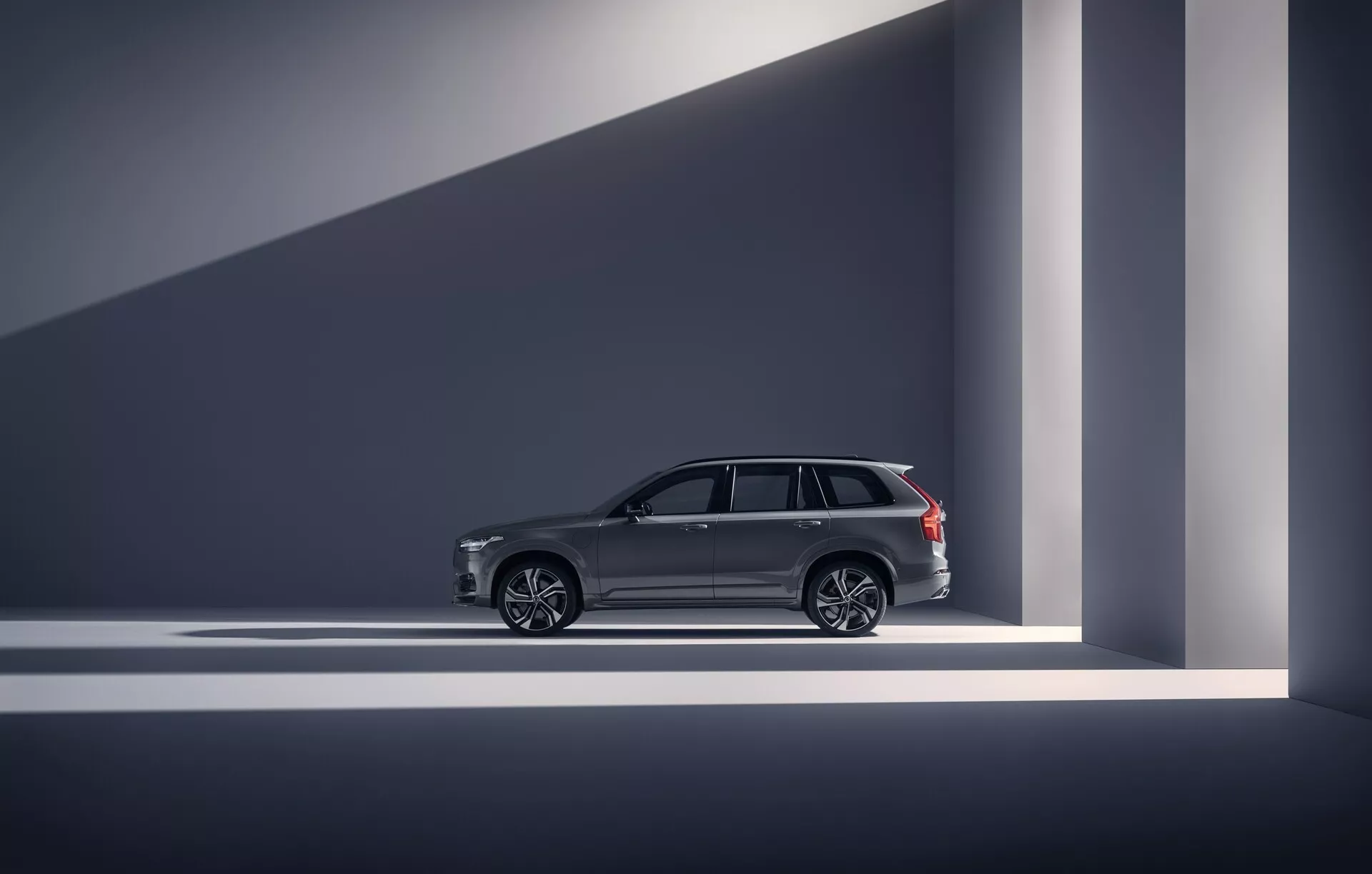 Profilo laterale di una Volvo XC90 2015 (2ª generazione) nella categoria SUV di lusso. La foto è scattata in uno studio o showroom al chiuso, mostrando le linee di design del veicolo, maniglie delle porte eleganti, grandi ruote in lega multiraggi e finiture cromate sui finestrini. Le barre sul tetto suggeriscono sia utilità che lusso. Il veicolo è ben illuminato con una luce drammatica che crea ombre geometriche, enfatizzando il suo aspetto moderno e di alta gamma. Le caratteristiche avanzate come l'accesso senza chiave sono indicate attraverso elementi di design.