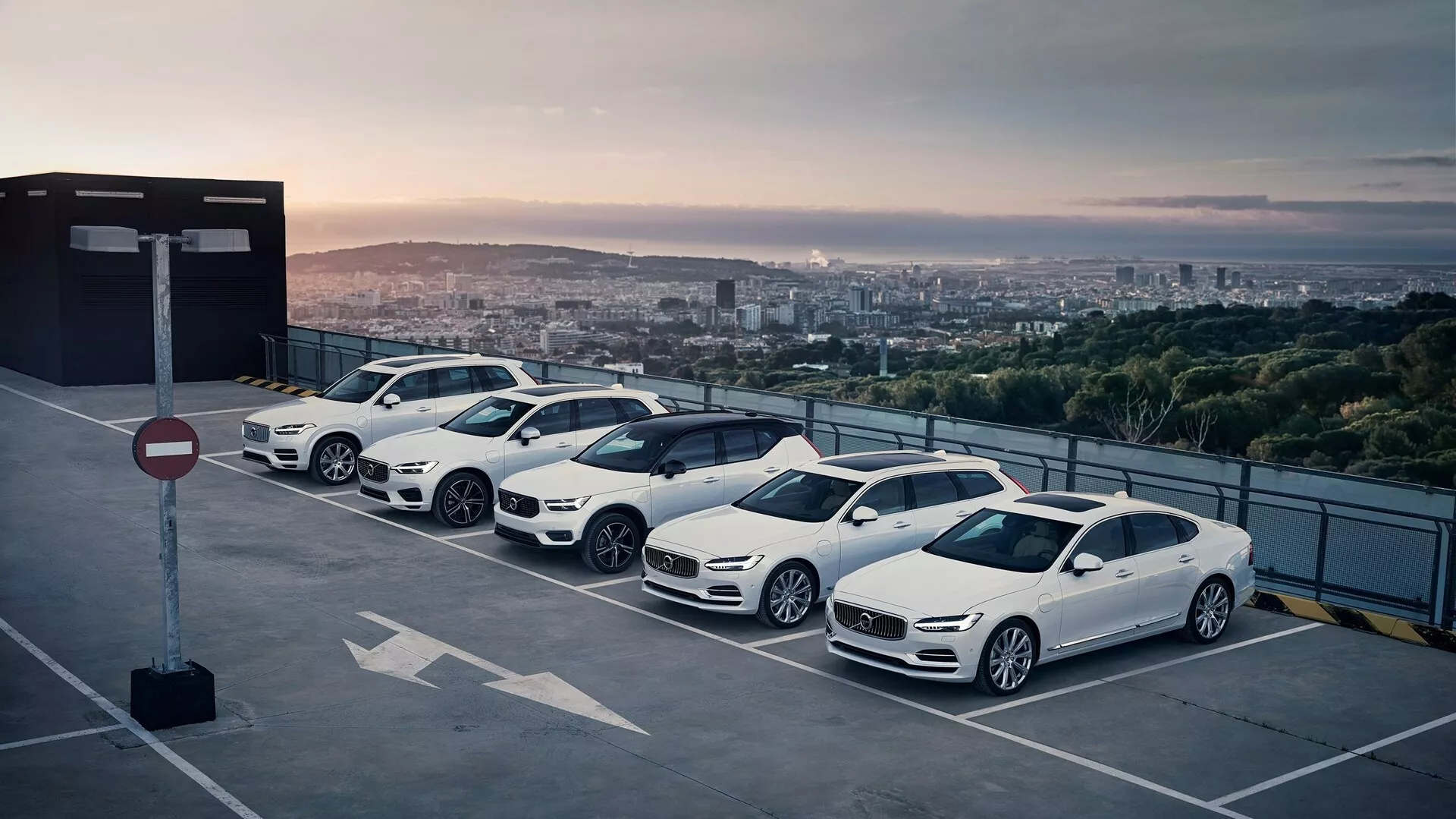 Vista elevata di quattro auto Volvo XC90 2015 (2ª generazione) parcheggiate in diagonale su un tetto durante l'alba o il tramonto, evidenziando i loro profili laterali destri su uno sfondo cittadino scenografico.