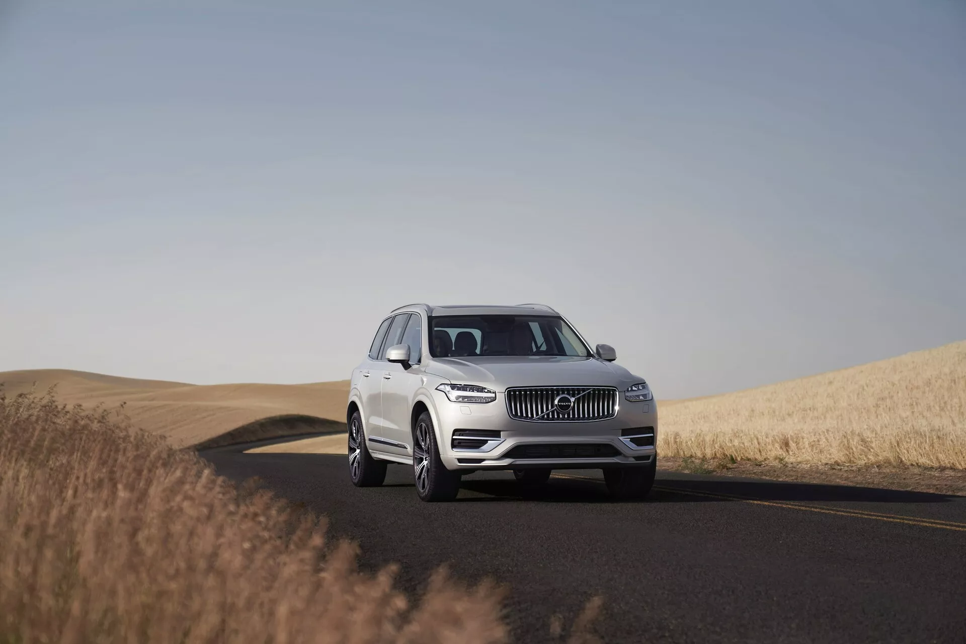 Un SUV Volvo XC90 bianco del 2015 su una strada a due corsie, catturato durante il giorno con un cielo limpido. L'immagine mostra il veicolo da un angolo basso a medio, evidenziando la sua presenza imponente e le dimensioni. Posizionato al centro del frame con una vista a tre quarti, la foto cattura dettagli di design come la griglia, i fari LED, l'emblema e il paraurti anteriore, indicanti sistemi avanzati di assistenza alla guida. Le ruote, le leghe e, eventualmente, la dimensione degli pneumatici sono chiaramente visibili. L'ambiente sereno presenta una strada che curva a destra e campi di grano dorati, evidenziando l'idoneità del veicolo per lunghi viaggi in vari paesaggi.