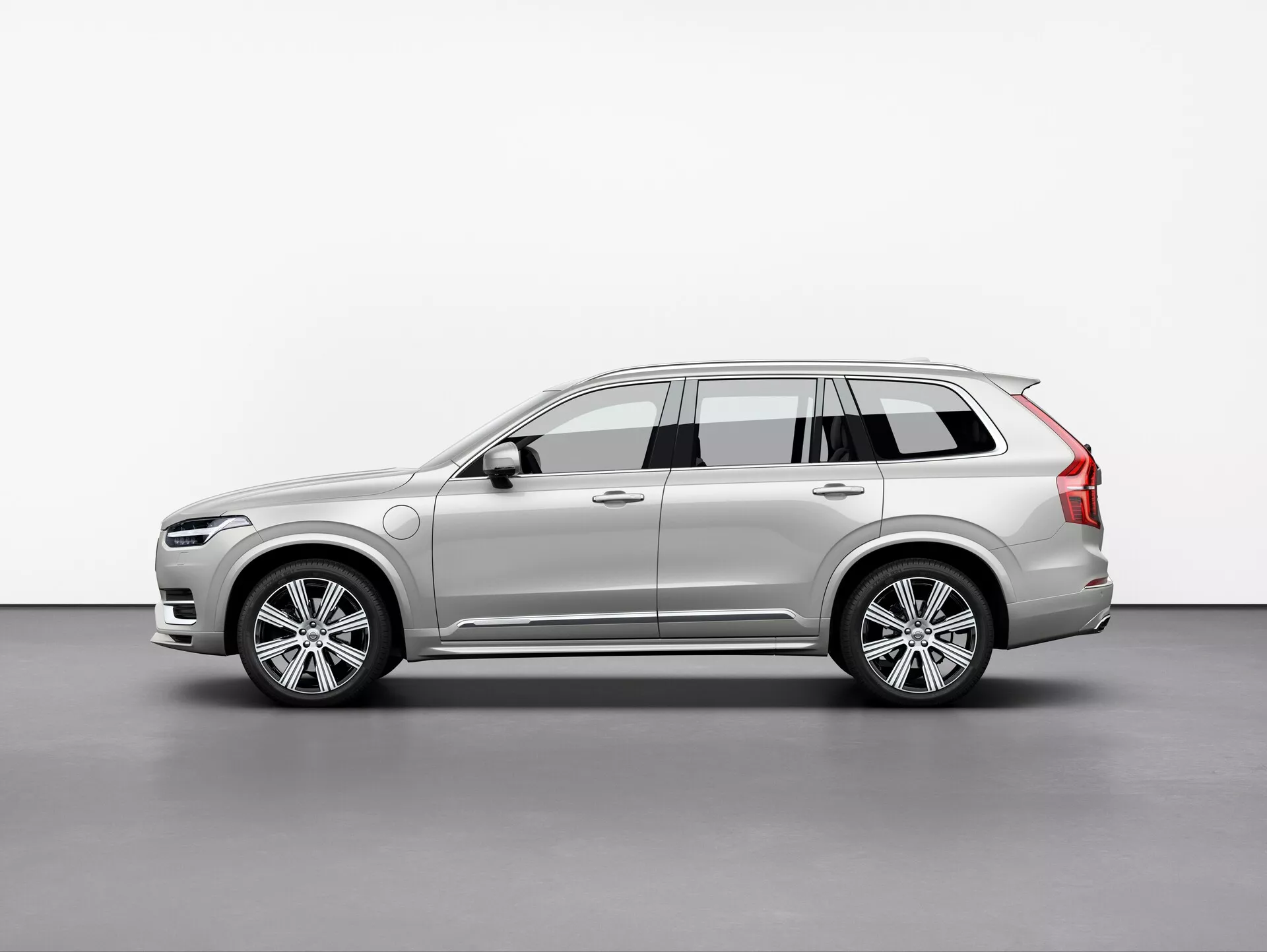 Immagine del SUV Volvo XC90 2015, fotografato da una vista a tre quarti con un leggero angolo. Questa vista evidenzia i profili anteriore e laterale, mettendo in mostra il design elegante e moderno dell'auto, i fari LED intricati, la griglia frontale parzialmente visibile, le ruote in lega di alluminio a più razze, gli specchietti laterali con indicatori di direzione integrati, le barre sul tetto, le linee dinamiche della carrozzeria, la vernice e il colore. L'illuminazione soffusa nell'ambito dello studio riflette accuratamente i contorni e i dettagli di superficie del veicolo.