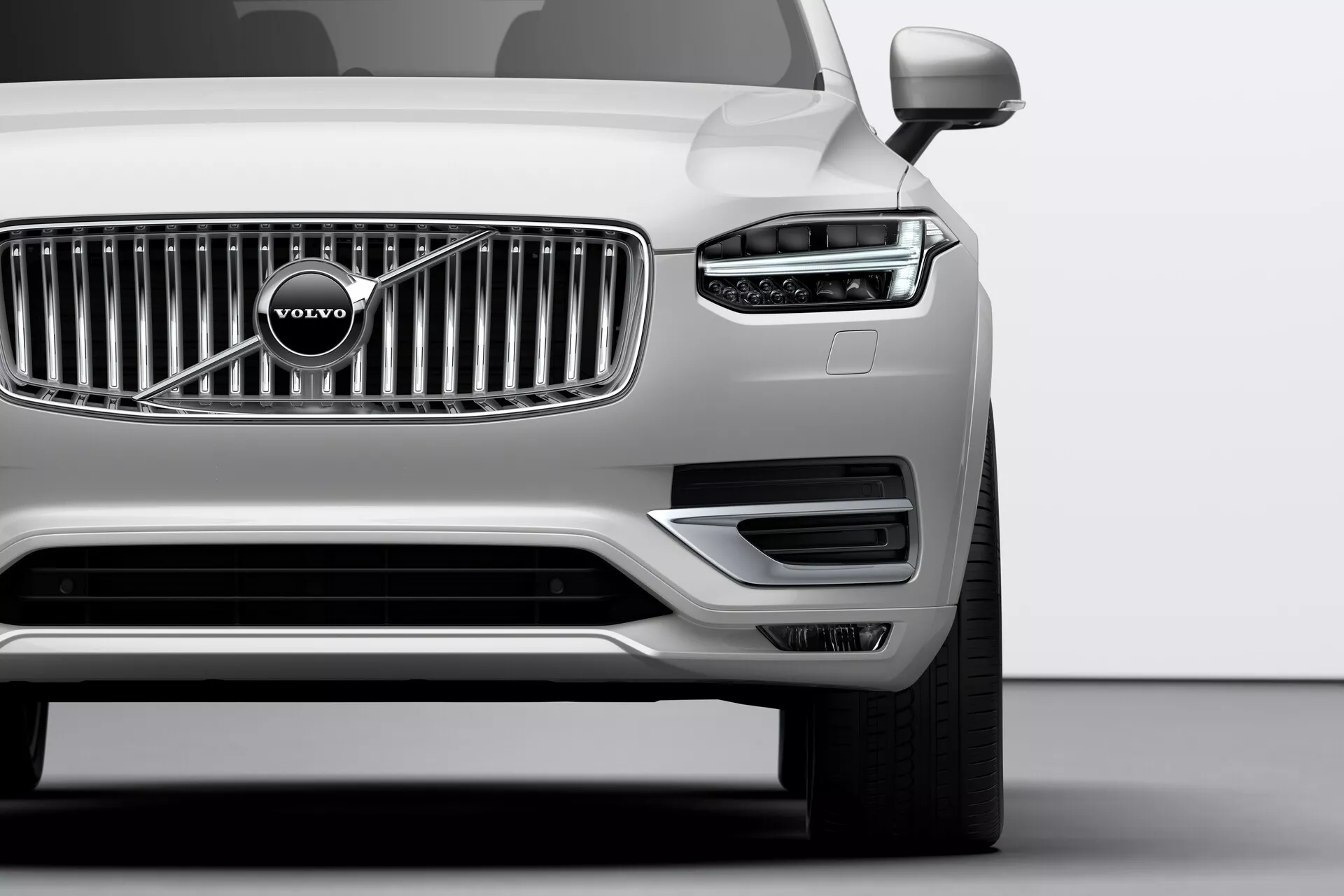 Immagine ravvicinata del quarto anteriore di una Volvo XC90 del 2015 (2ª generazione) argento, scattata da un angolo basso. Evidenzia la prominente griglia cromata con l'emblematico logo Volvo, i fari eleganti con luci diurne a LED a forma di T, noti come 'Martello di Thor', e i sensori di parcheggio ben integrati nel paraurti anteriore. La parte inferiore del paraurti include prese d'aria e alloggiamenti per i fendinebbia incorniciati da finiture argento. Gli specchietti esterni visibili suggeriscono funzionalità tecnologiche avanzate come indicatori di direzione o sistemi di avviso per l'angolo cieco.