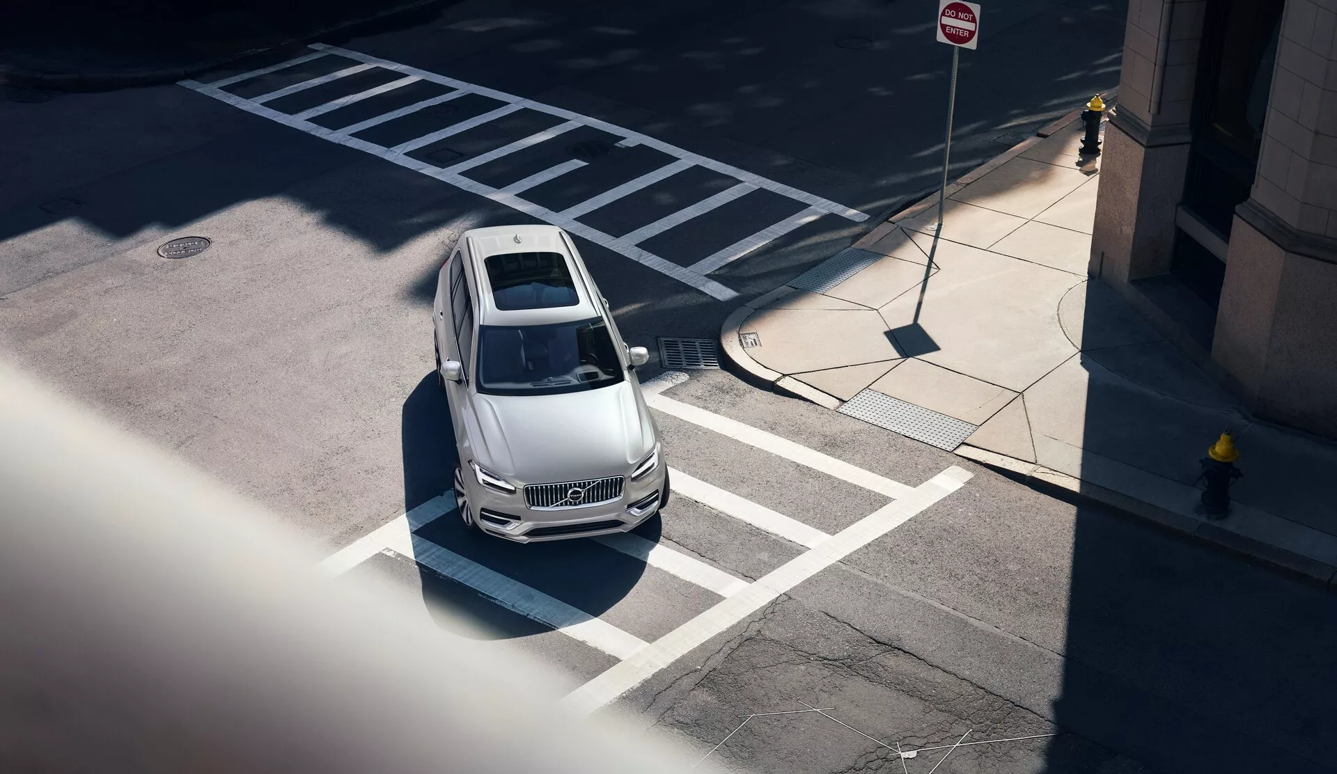 Vista dall'alto di una Volvo XC90 del 2015 (2ª generazione) da un alto angolo, vista diagonalmente dall'angolo anteriore sinistro; che mostra il cappuccio di colore chiaro, il tetto e una parte del lato del conducente. L'auto, correttamente parcheggiata in uno spazio contrassegnato vicino a un passaggio pedonale, presenta contorni di design indicativi di uno styling aerodinamico, un parabrezza chiaro rivela un interno parzialmente visibile, un tetto apribile visibile e linee laterali nitide. Il contesto urbano è enfatizzato dalle vicine segnaletiche del parcheggio e del traffico, con ombre pronunciate che suggeriscono un'illuminazione di mezzogiorno. Il veicolo lascia anche intuire caratteristiche tecnologiche moderne senza sensori o telecamere visibili.