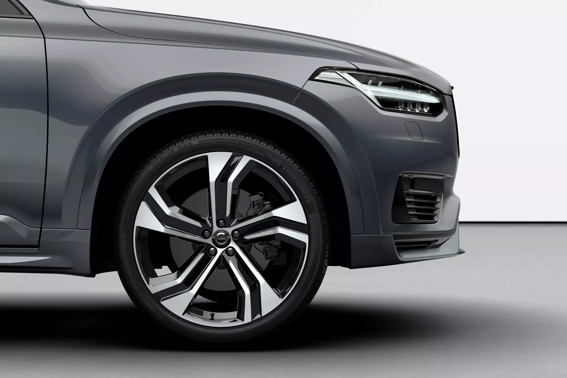 Ripresa angolare bassa della ruota anteriore lato conducente e delle sezioni adiacenti di una Volvo XC90 del 2015, che mostra il design moderno del cerchio in lega con più raggi, il pneumatico sportivo a profilo ribassato, parte del parafango anteriore con linee aerodinamiche, il design elegante del faro, il paraurti anteriore con motivo della griglia orizzontale, un possibile fendinebbia o presa d'aria sotto il faro e la finitura della vernice metallizzata, tutto indicativo di un veicolo di lusso o di livello superiore.