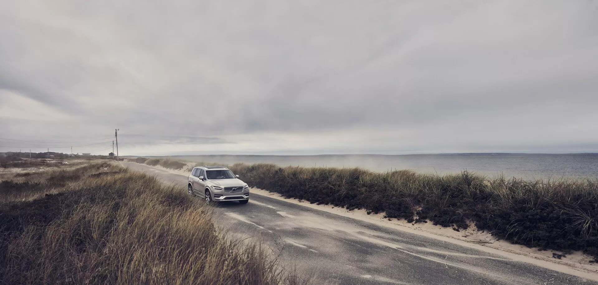 Foto grandangolare di una Volvo XC90 del 2015 (2a generazione) che guida su una stretta strada costiera. L'auto si avvicina alla telecamera, mettendo in evidenza la griglia anteriore, i fari e il paraurti anteriore. Con un cielo nuvoloso e l'oceano sullo sfondo, il colore chiaro e metallico del SUV risalta. Questa scena enfatizza la combinazione di lusso e avventura del veicolo, suggerendo funzionalità tecnologiche e di sicurezza avanzate per le condizioni di guida costiere.