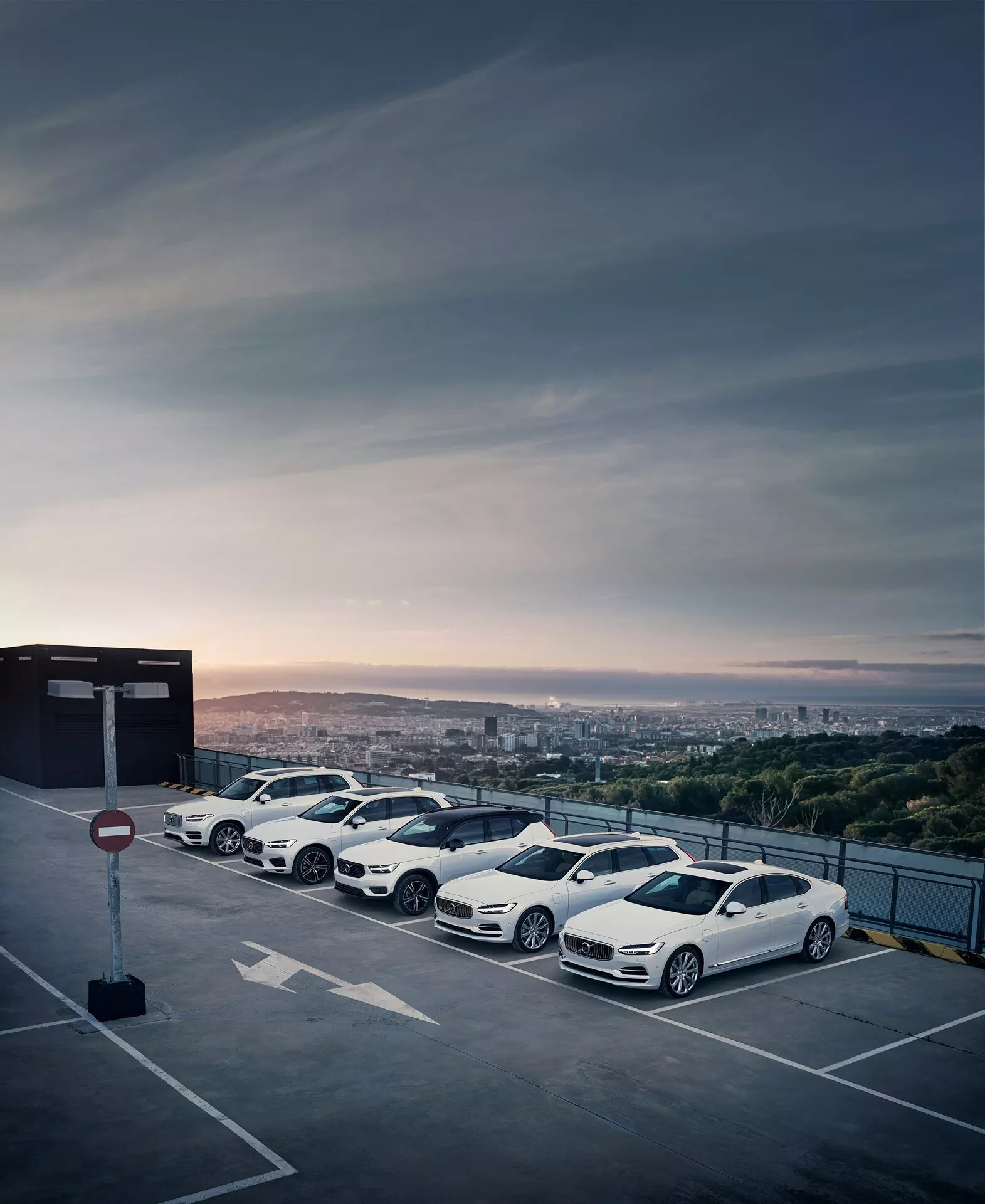 Schieramento di quattro auto moderne e lussuose Volvo XC90 2015 (2ª generazione) parcheggiate al piano superiore di una struttura di parcheggio elevata durante l'alba o il tramonto con uno skyline urbano sullo sfondo. Le berline bianche presentano design aerodinamici, cerchi in lega a più razze, specchietti laterali aerodinamici e vetri oscurati, mettendo in mostra i loro contorni eleganti e un'estetica dinamica.