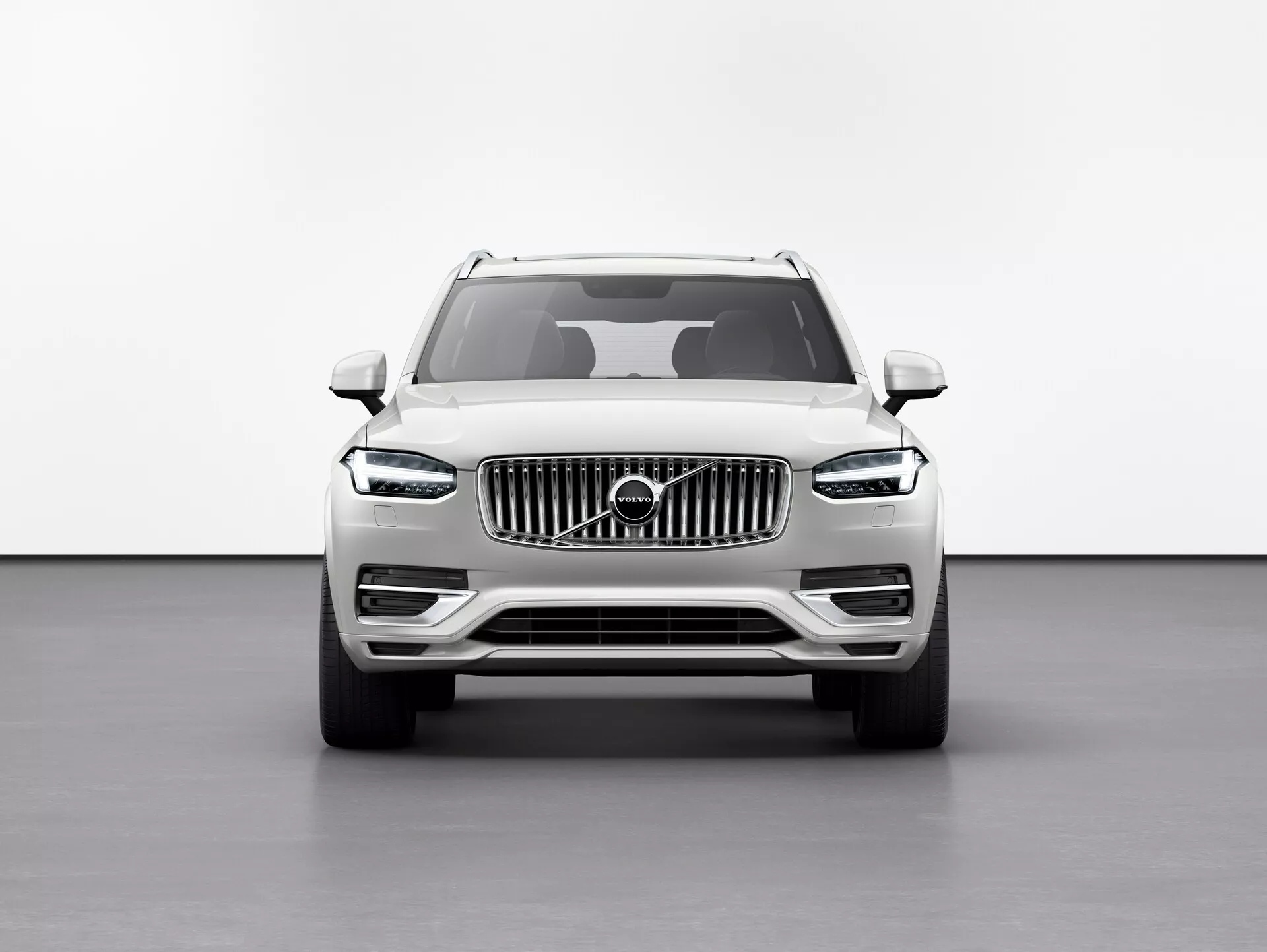 Vista frontale del SUV Volvo XC90 2015 (2ª generazione) su sfondo bianco in un ambiente da studio, evidenziando l'altezza da terra elevata, la grande griglia a barre verticali con emblema Volvo, i raffinati fari a LED 'martello di Thor', linee definite del cofano, fendinebbia nel paraurti inferiore e specchietti laterali con indicatori di direzione integrati.