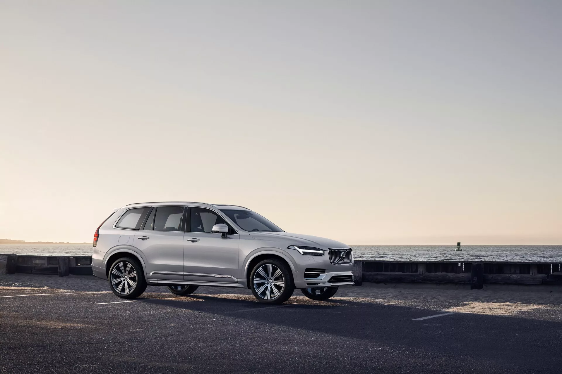 Profilo laterale di un SUV Volvo XC90 2015 bianco su una strada costiera, con dettagli di design visibili tra cui il design delle ruote, i pannelli delle portiere, gli specchietti laterali, le barre del tetto e la configurazione dei finestrini accentuati dalla morbida luce dell'alba o del tramonto.