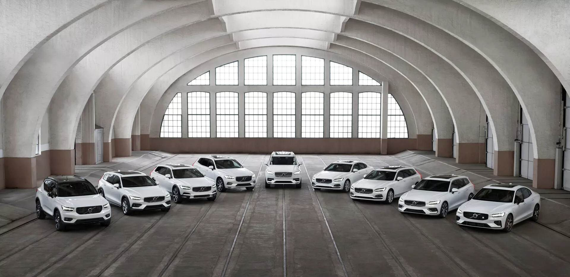 Una fila di otto auto Volvo XC90 2015 di seconda generazione, bianche, posizionate all'interno di un edificio spazioso e architettonicamente classico, con grandi finestre arcuate e un soffitto curvo. I veicoli sono orientati verso la telecamera ad angoli diversi, mostrando le loro parti frontali e caratteristiche di design, inclusi fari a LED, grandi ruote in lega e pannelli della carrozzeria scolpiti. L'illuminazione mette in risalto gli elementi di design delle auto, mentre il pavimento, segnato da tracce di pneumatici, suggerisce l'uso dello spazio per il parcheggio dei veicoli.