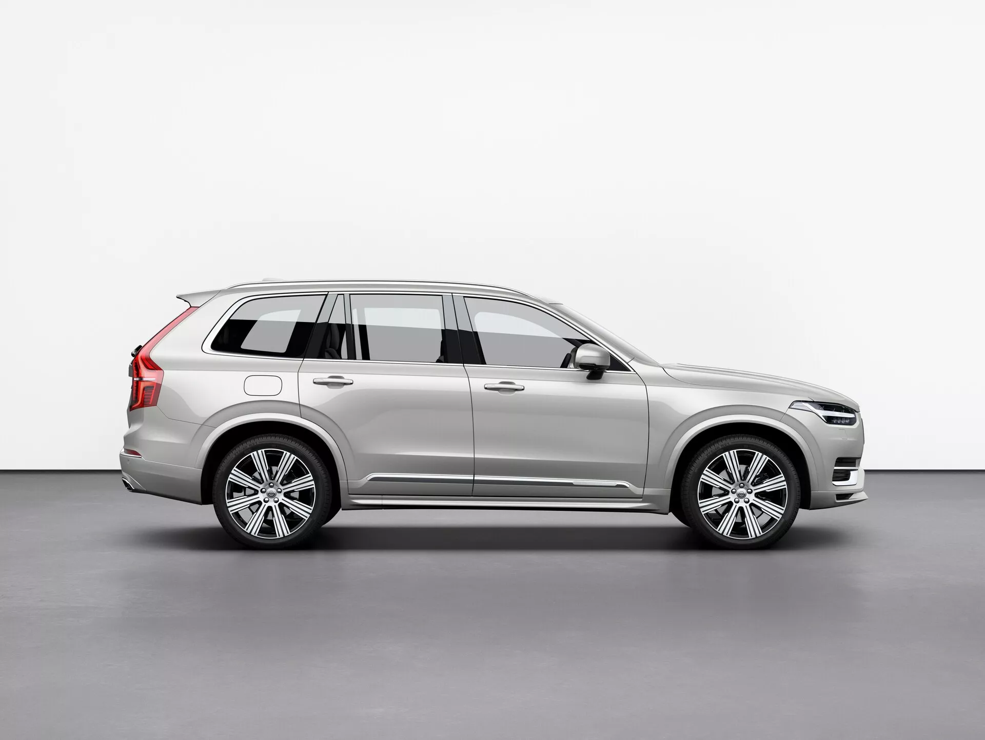 Immagine di profilo laterale di un SUV Volvo XC90 2015 (2ª generazione) argento, che mostra le sue ruote in lega, le barre del tetto, lo specchio laterale con indicatore di direzione integrato e le linee dettagliate della carrozzeria, posizionata centralmente su uno sfondo semplice.