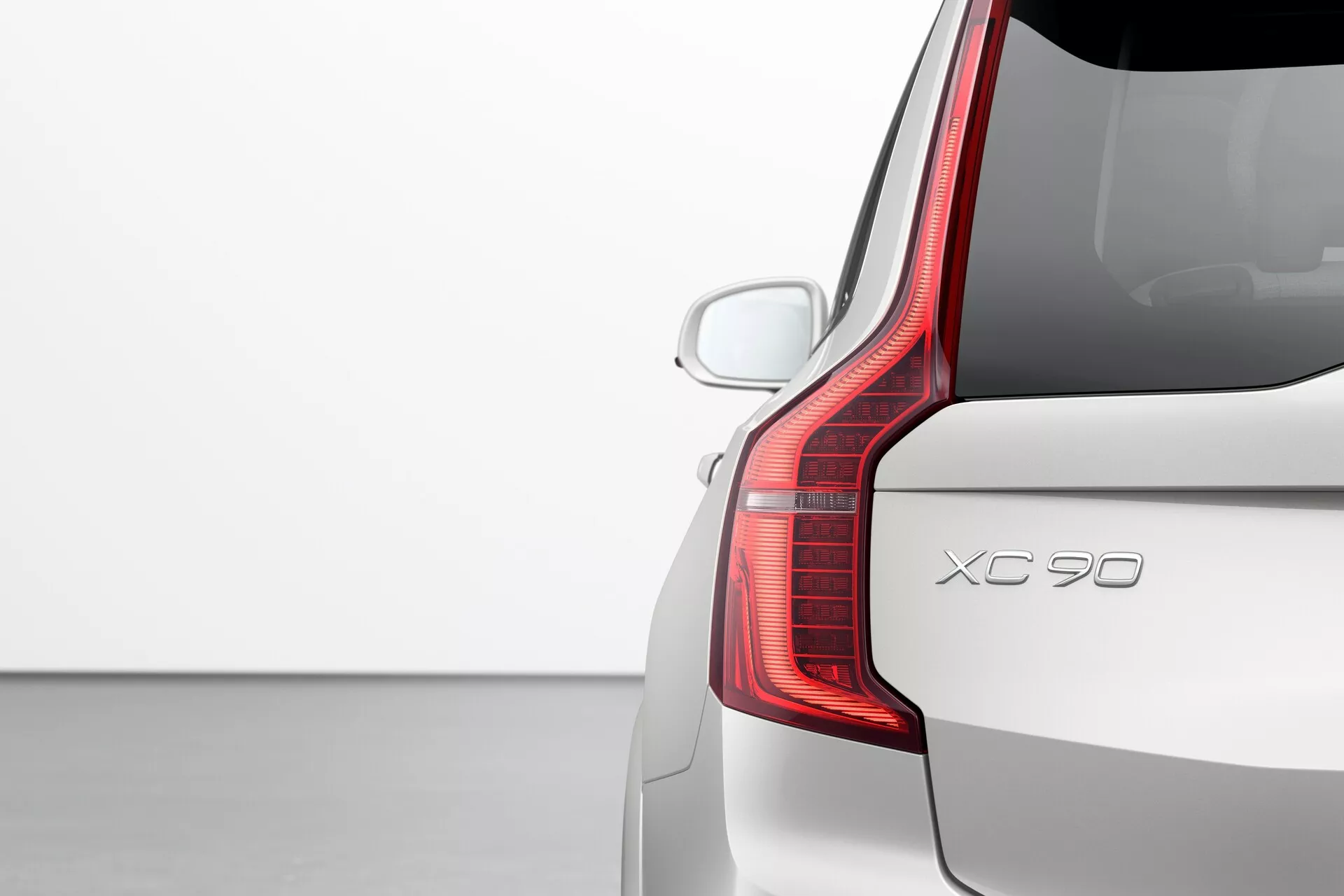 Vista dettagliata del quarto posteriore lato passeggero di una Volvo XC90 2015 (2ª generazione), che mostra il distintivo design delle luci posteriori a LED, il portellone e il paraurti posteriore. L'angolo posteriore a tre quarti enfatizza l'estensione verticale della luce posteriore e la sua forma unica, che si curva in avanti verso il lato. L'immagine evidenzia anche il parabrezza posteriore con lo spoiler aerodinamico sul tetto, un sensore di parcheggio posteriore visibile e la vernice di colore solido in un ambiente di studio semplice, focalizzandosi sul design e le caratteristiche dell'auto.