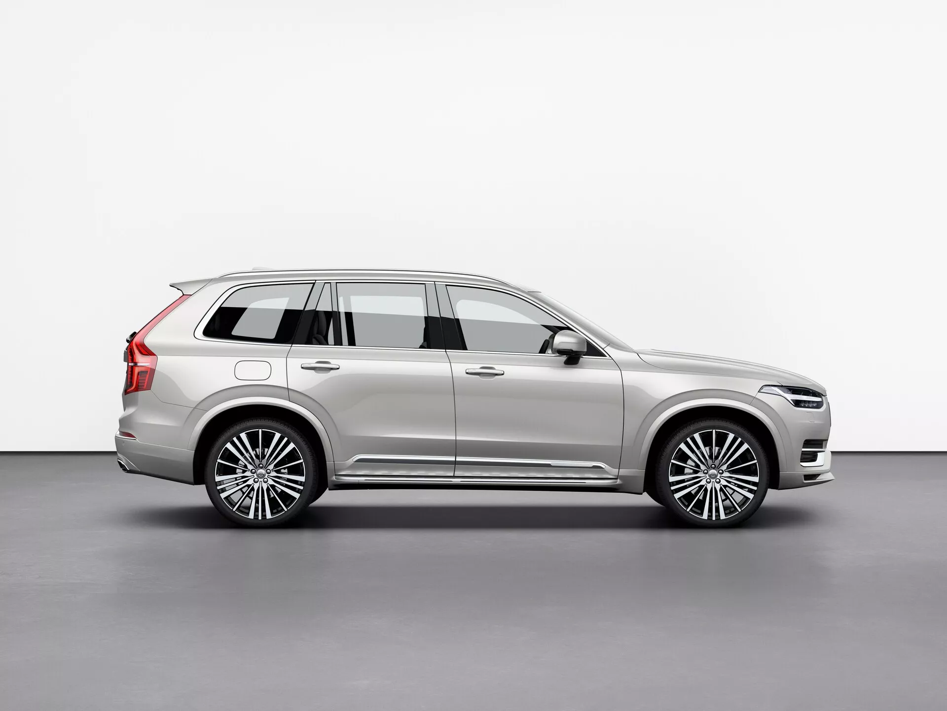 Profilo laterale della Volvo XC90 2015, un SUV di lusso di grandi dimensioni, catturato in uno studio con sfondo bianco. L'immagine mostra il lungo passo del veicolo, le quattro porte, il portellone posteriore, i cerchi in lega multirazze, le finiture cromate, la verniciatura metallizzata e gli elementi di design predominanti come gli specchietti laterali e le barre del tetto. La scena accuratamente illuminata mette in risalto le linee e le curve della carrozzeria, riflettendo la sua natura premium. Il veicolo è posizionato per fornire una vista chiara della lunghezza, altezza e postura complessiva, indicativa di un SUV con capacità a tre file per passeggeri e carico.
