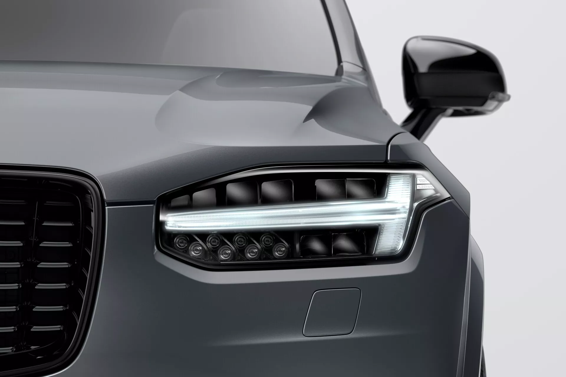 Primo piano del frontale di una Volvo XC90 2015 (2ª generazione), con particolare attenzione al faro sinistro con tecnologia moderna a LED o laser, doppi elementi di illuminazione, DRL eleganti e diverse lenti del proiettore per una visibilità migliorata. L'immagine enfatizza la grande griglia esteticamente progettata, lo specchio laterale aerodinamico in tinta carrozzeria e il possibile sensore ADAS sotto il faro. La finitura grigio metallizzato scuro liscia e riflettente mette in risalto le linee eleganti ma muscolose del cofano e del parafango, dimostrando una verniciatura di alta qualità e un design veicolare all'avanguardia.