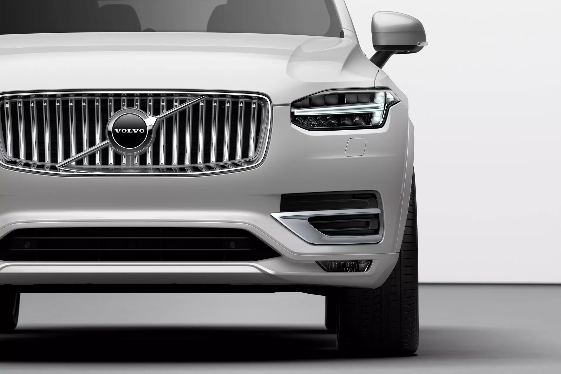 Immagine a bassa angolazione del quarto anteriore sinistro di una Volvo XC90 argento del 2015 (2ª generazione) in uno studio, che mostra il distintivo faro LED a martello di Thor, la griglia cromata a listelli verticali con il logo Volvo, il paraurti anteriore minimalista con prese d'aria, lo specchietto laterale sinistro, la ruota in lega a più razze e la finitura metallica impeccabile, illustrando la tecnologia avanzata dell'illuminazione, lo stile del marchio e le caratteristiche aerodinamiche.
