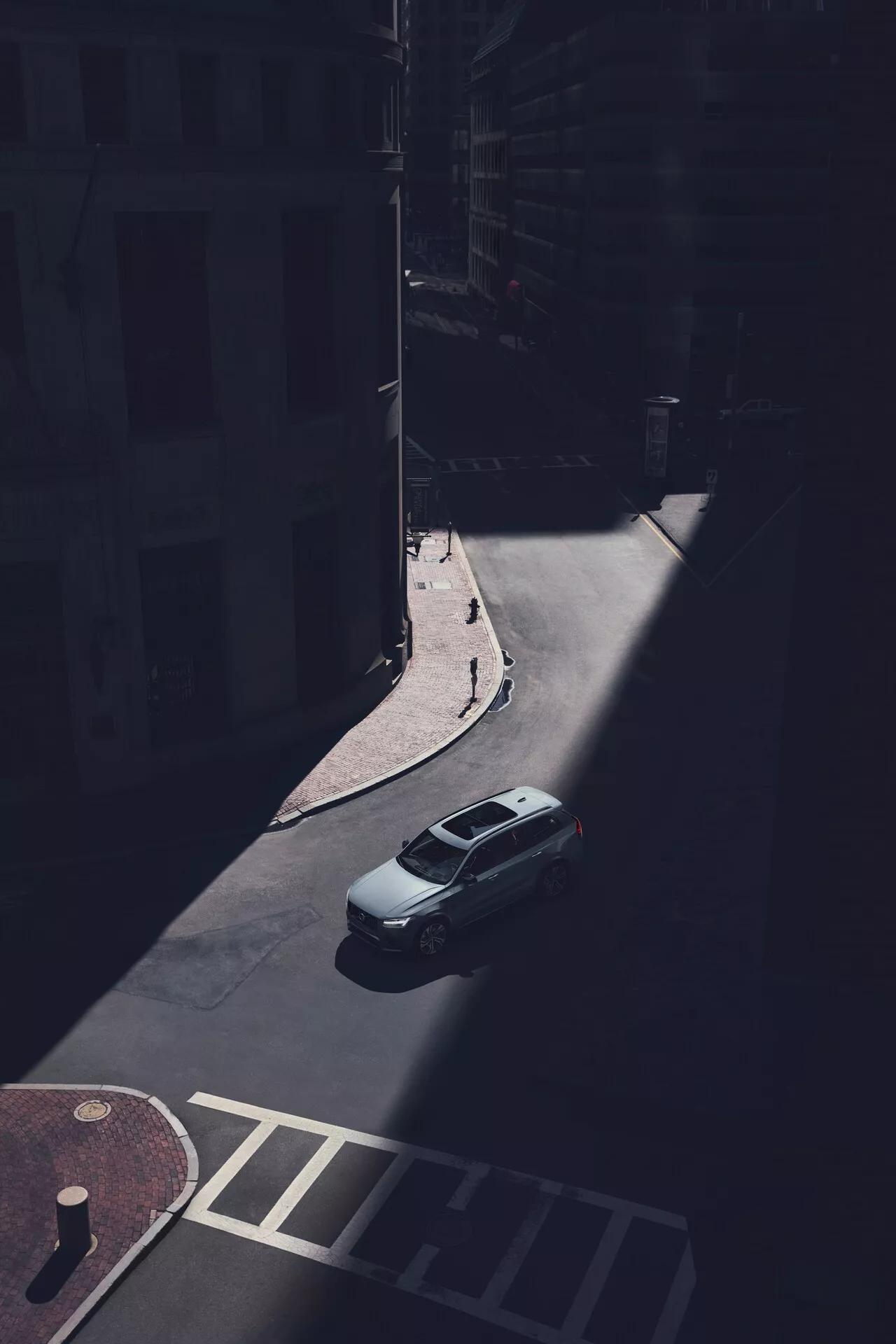 Vista aerea della berlina Volvo XC90 2015 (2ª generazione) parcheggiata su una strada urbana ombreggiata, catturata da un punto di vista elevato guardando verso il basso ad un angolo obliquo. Il bagagliaio, il tetto e il parabrezza posteriore dell'auto sono chiaramente visibili, con la parte posteriore del veicolo illuminata dalla luce solare. L'immagine cattura il coperchio del bagagliaio posteriore, la forma del lunotto posteriore e la parte superiore del bagagliaio, mentre le ombre oscurano i dettagli specifici del design, come i fanali posteriori e il paraurti. La linea del tetto aerodinamica è evidente, senza portapacchi o tetti solari visibili. L'auto è parcheggiata accanto a un marciapiede vicino alle facciate degli edifici e a un attraversamento pedonale, enfatizzando l'isolamento del veicolo nell'ambiente urbano ombreggiato.