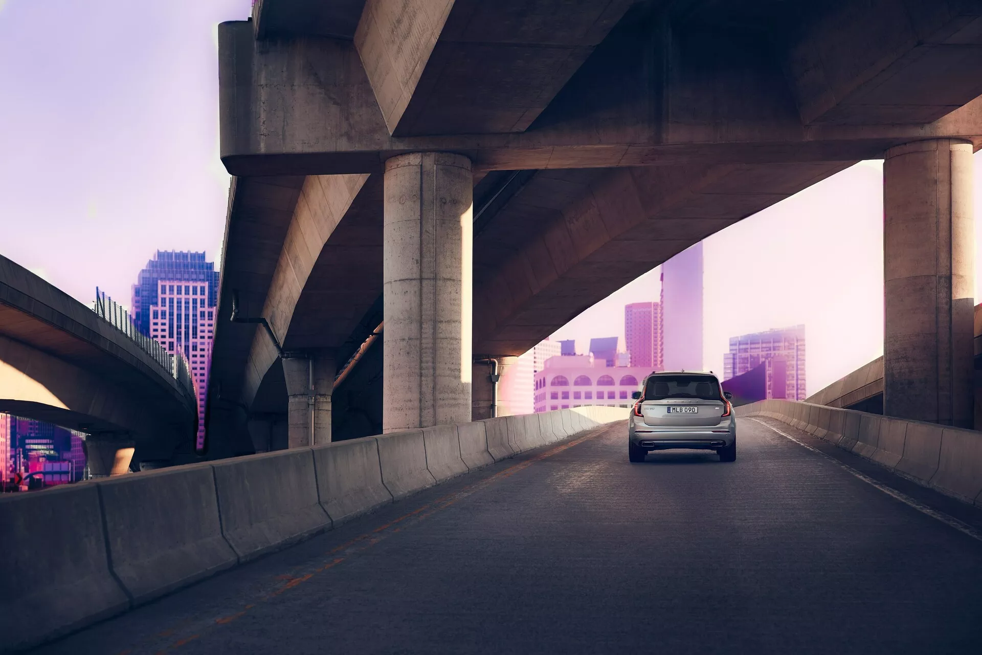 Vista tre quarti posteriore di una Volvo XC90 di seconda generazione del 2015 su un'autostrada sopraelevata, contro un paesaggio urbano leggermente illuminato di rosa-viola all'alba o al tramonto. Le luci dei freni dell'auto sono accese, suggerendo una decelerazione. Il SUV presenta una linea del tetto alta, parte posteriore con portellone e design aerodinamico per efficienza e stile, catturato in luce naturale diffusa.