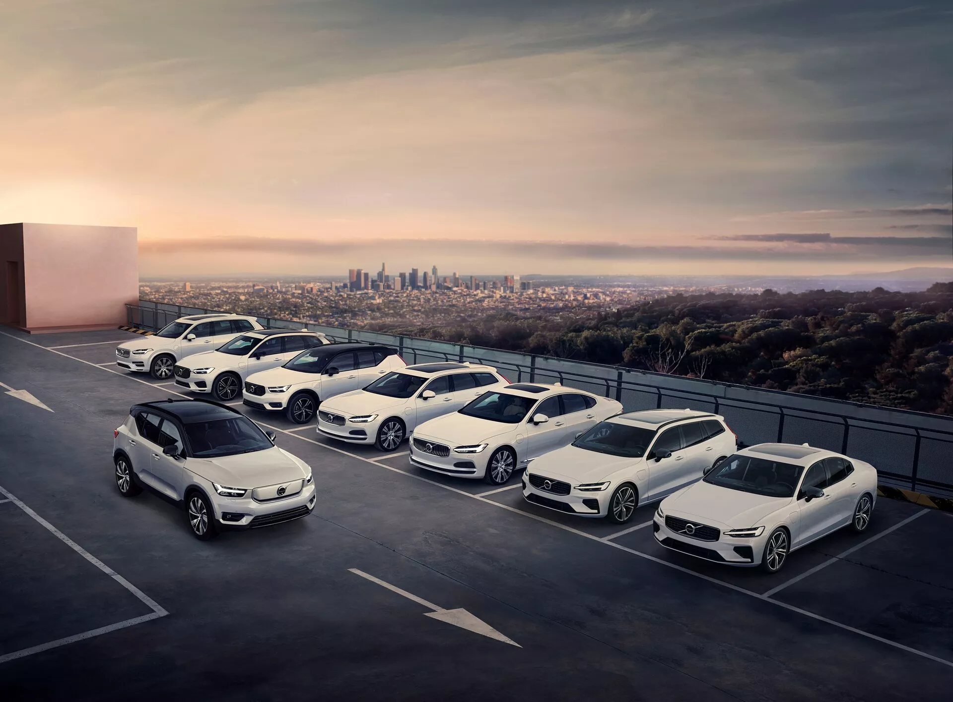 Vista elevata di cinque auto parcheggiate in diagonale sul livello superiore di un parcheggio multipiano, con lo skyline della città sullo sfondo. I veicoli includono una varietà di SUV compatti, berline e modelli sportivi, tutti con elementi di design moderni come fari a LED e cerchi in lega. Il contesto, catturato al crepuscolo, aggiunge un'atmosfera premium e serena alla scena.