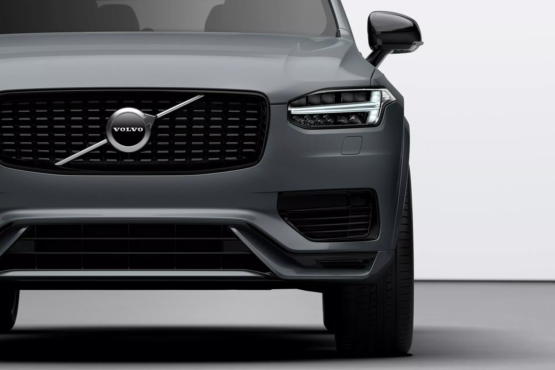 Vista ravvicinata dell'angolo frontale della Volvo XC90 2015 (seconda generazione) che si concentra sul lato sinistro, mettendo in evidenza la griglia distintiva, il grande emblema Volvo e il design del faro a LED 'Martello di Thor'. L'immagine include il faro, la ruota anteriore, parte della porta del conducente, sensori visibili sul paraurti inferiore e lo specchietto laterale con possibile tecnologia di monitoraggio dell'angolo cieco.