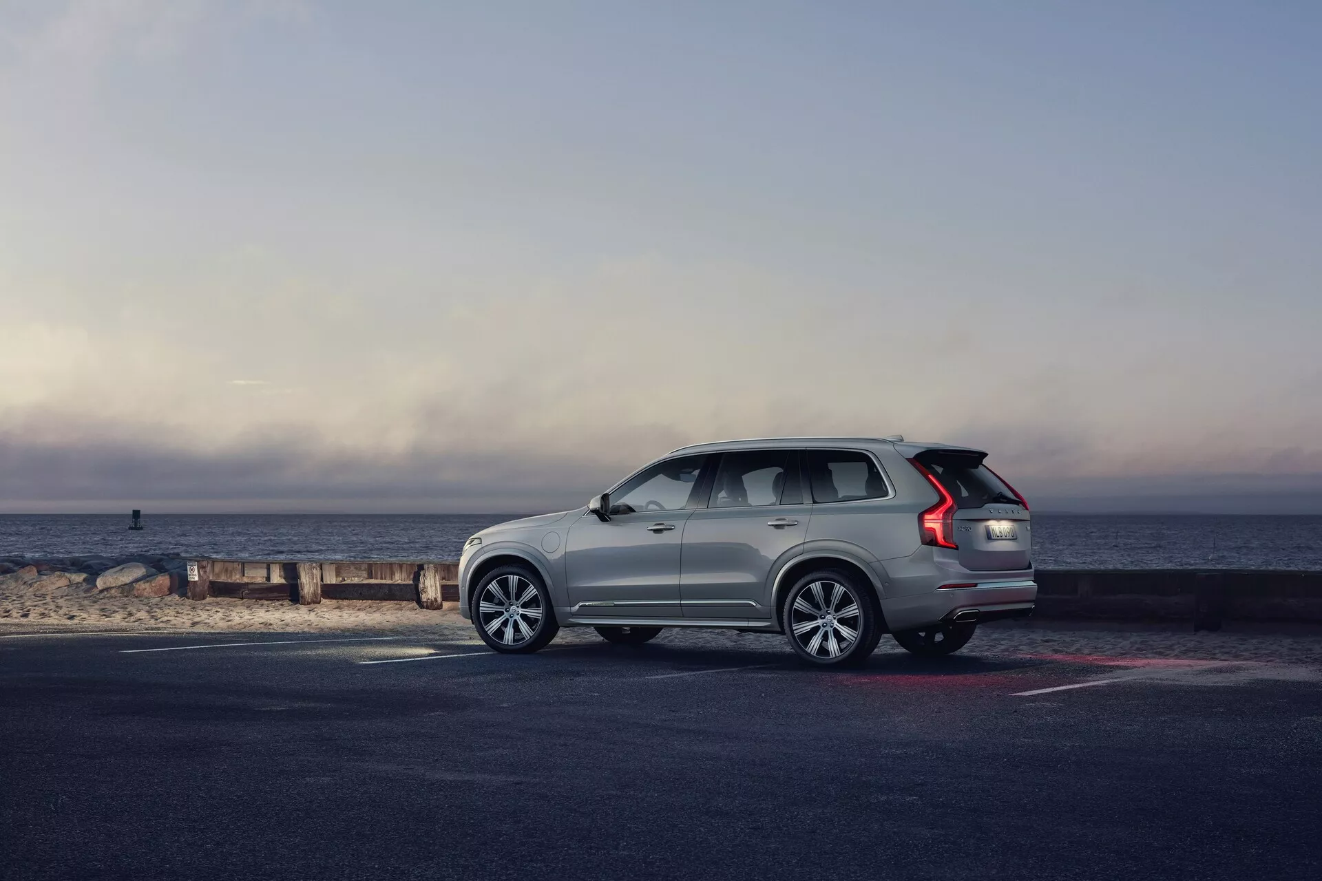 Vista di profilo di un SUV Volvo XC90 2015 (2ª generazione) parcheggiato sulla riva, con il veicolo rivolto verso destra. L'angolazione enfatizza il lato e il retro, mostrando elementi di design come le ruote in lega multirazze, le luci posteriori a LED allungate, le barre sul tetto, la linea dei finestrini che sale verso il posteriore e le maniglie delle porte a filo. Il SUV di colore chiaro risalta sul pavimento più scuro e sulla spiaggia, bagnati dalla luce naturale del mattino presto o del tardo pomeriggio, con uno sfondo panoramico dell'orizzonte dove il cielo incontra il mare. La composizione generale evoca un senso di avventura, lusso e libertà.