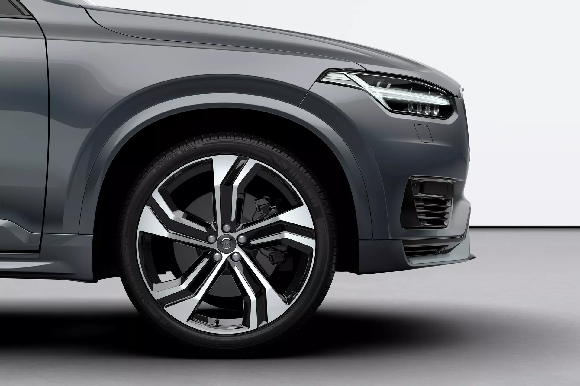 Vista ravvicinata e angolazione bassa della ruota anteriore e di una parte del quarto anteriore di una Volvo XC90 del 2015. L'immagine cattura il cerchio in lega multirazza, parte del paraurti anteriore con fendinebbia integrato o presa d'aria, e un'unità faro con elementi di design moderni. La finitura della vernice metallizzata e il design scolpito del parafango sottolineano le caratteristiche premium del SUV, enfatizzando l'alto arco della ruota e il robusto pneumatico. L'illuminazione in studio evidenzia gli attributi lussuosi dell'auto.