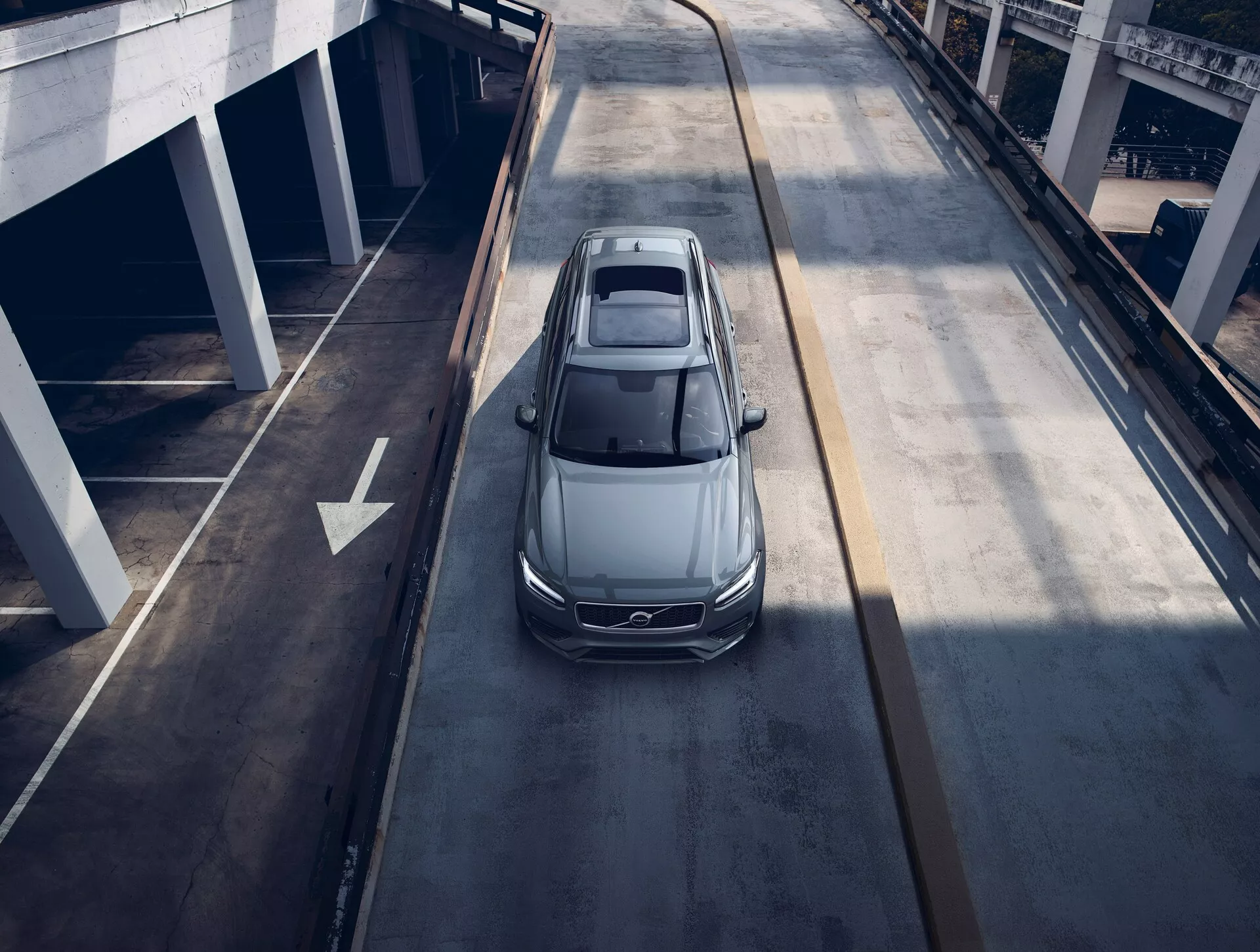 Vista dall'alto di una berlina Volvo XC90 2015 grigia o argento che viaggia su una strada urbana a più livelli, mostrante il tetto panoramico, il design aerodinamico, i fari LED eleganti e i grandi cerchi in lega, suggerendo un veicolo moderno e di lusso.
