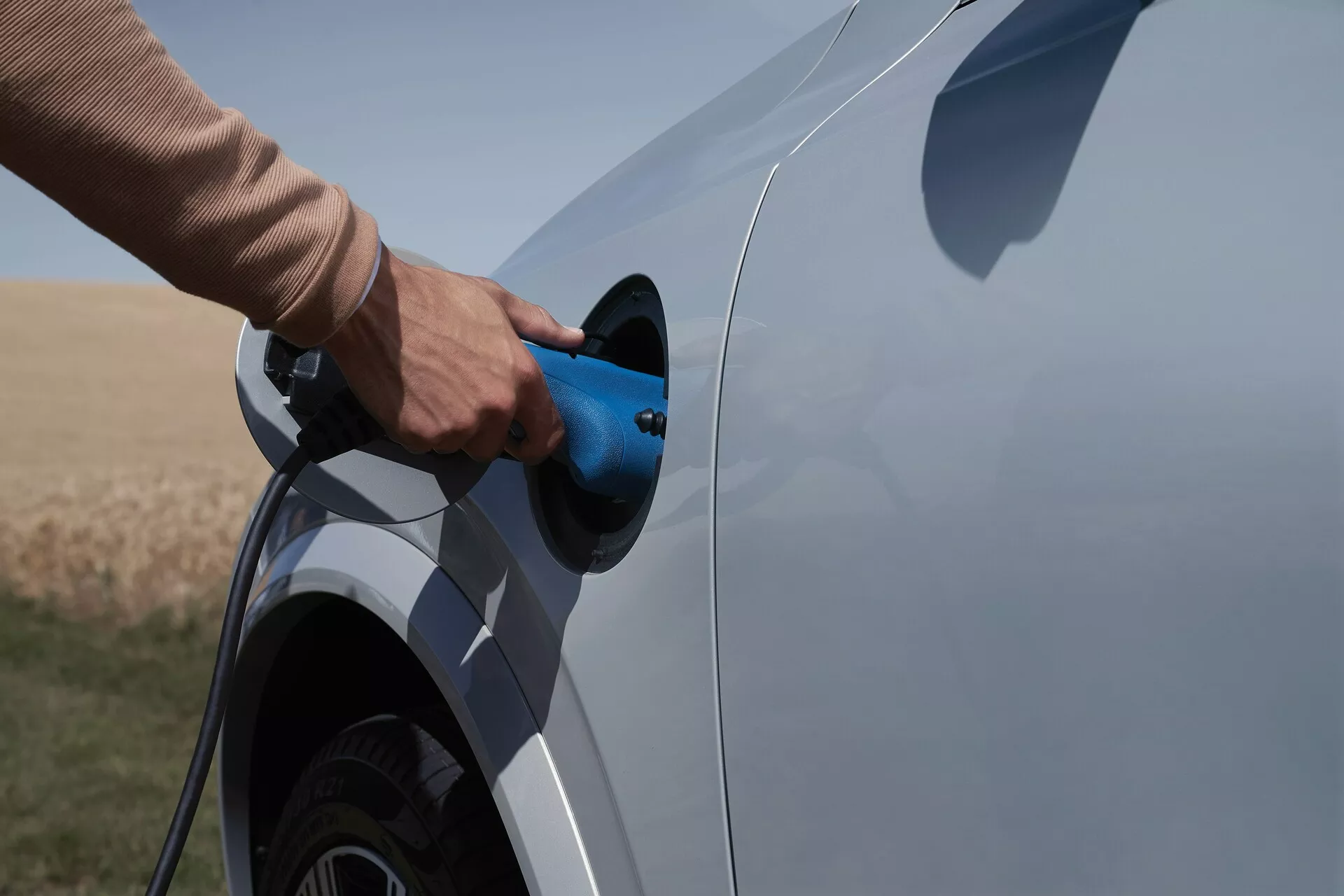Vista laterale di un veicolo elettrico o ibrido plug-in Volvo XC90 2015 di seconda generazione durante il processo di ricarica. La fotografia cattura il lato del veicolo con particolare attenzione al porto di ricarica e alla spina, mostrando parzialmente la ruota posteriore e un segmento del fondo dell'auto. Cavo di ricarica blu in fase di inserimento, sfondo di area naturale aperta, indicativo di un ambiente rurale.