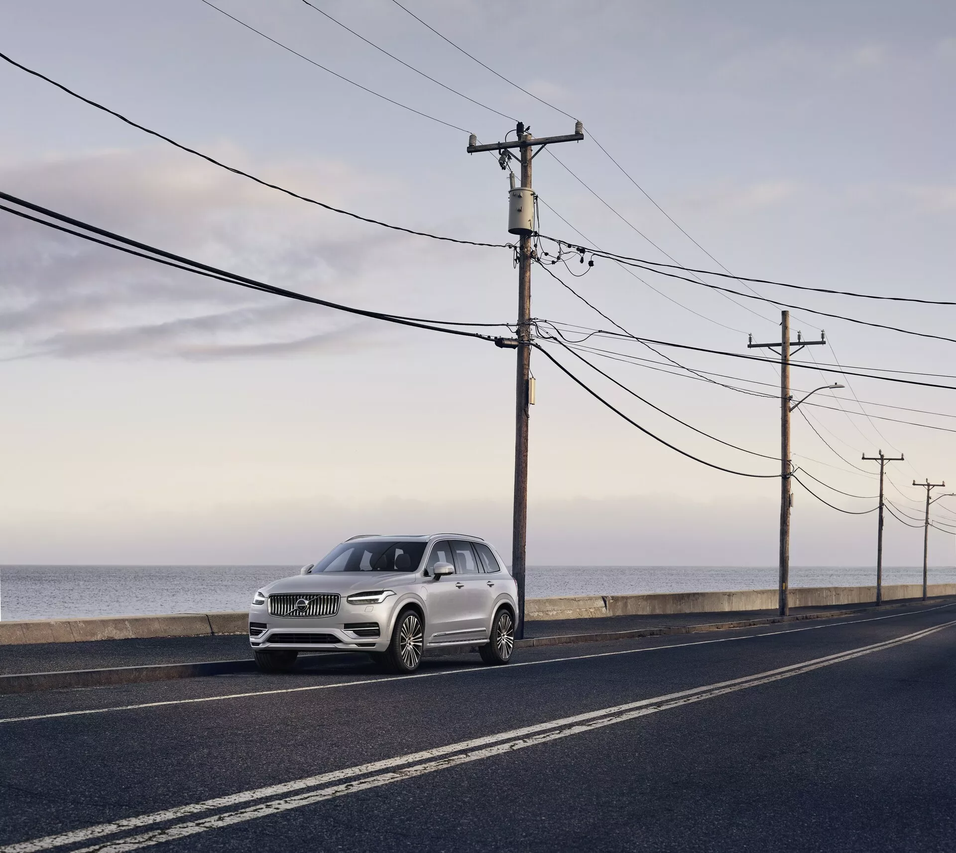 Volvo XC90 di seconda generazione del 2015, SUV di lusso parcheggiato su una strada asfaltata parallela alla costa oceanica, catturato alla luce del giorno con luce diffusa e morbida e lunghe ombre. Vista da un'angolazione laterale anteriore, evidenziando gli elementi di design eleganti, inclusi i fari, la griglia e i contorni del cofano, colore argento chiaro o champagne pallido che contrasta con la strada scura e lo sfondo naturale.
