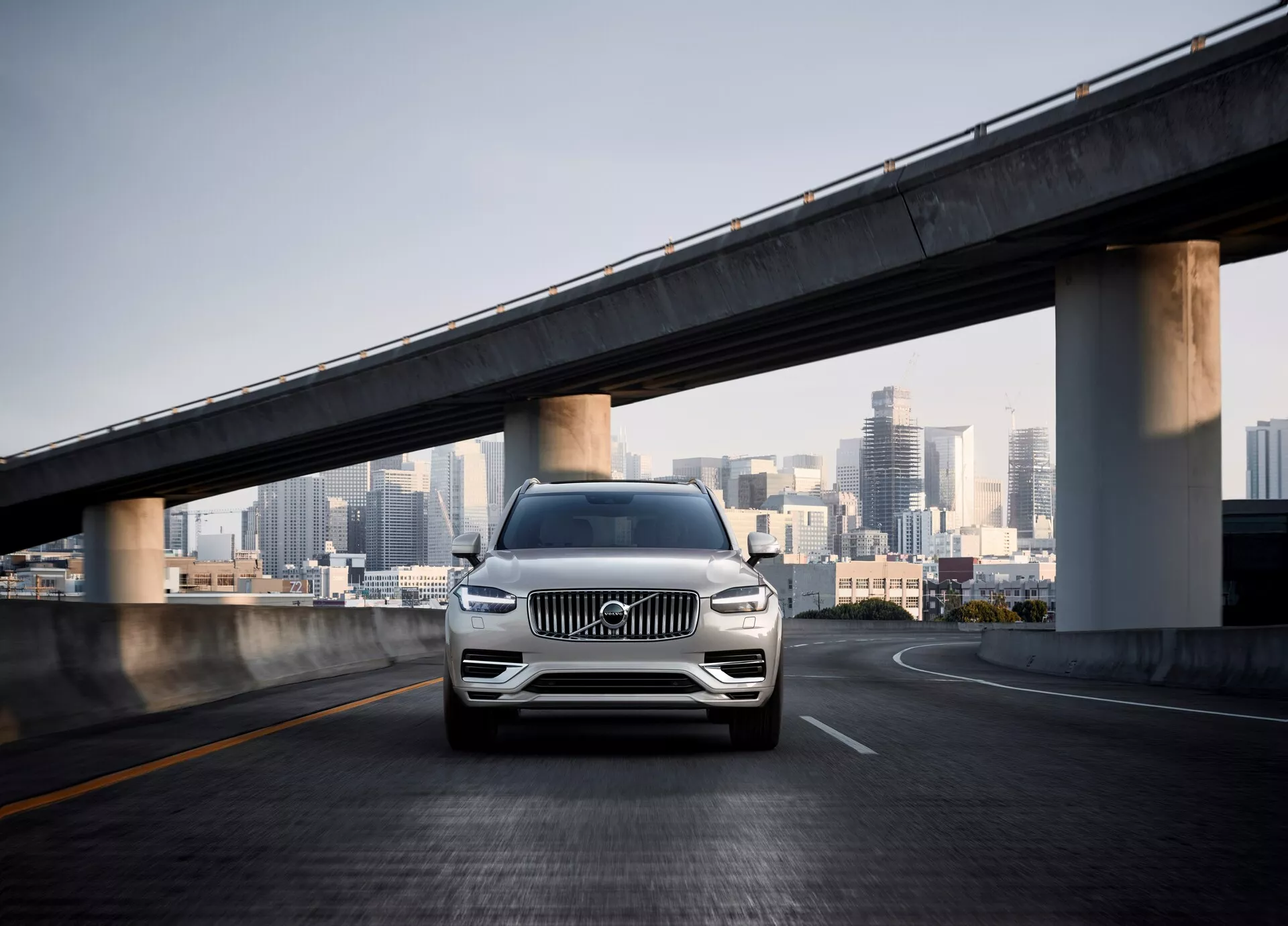 Una ripresa frontale del SUV Volvo XC90 di seconda generazione del 2015 su un cavalcavia vuoto, con lo skyline della città sullo sfondo. L'angolo della fotocamera è basso, mostrando la griglia anteriore, i fari LED, gli accenti cromati, le ruote in lega e un paraurti scolpito con sensori, indicando assistenza al parcheggio o sistema di evitamento delle collisioni. La vernice perlata o metallica dell'auto brilla alla luce del giorno, enfatizzando il suo lusso e design moderno in un ambiente urbano.