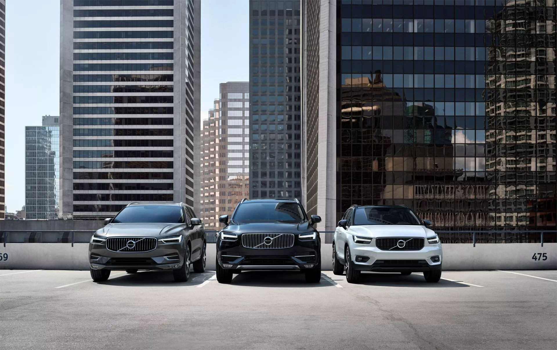 Tre auto di lusso Volvo XC90 2015 (2ª generazione) parcheggiate in parallelo, rivolte verso la fotocamera, con uno skyline urbano moderno sullo sfondo. La prospettiva frontale dei veicoli mostra fari a LED eleganti, grandi griglie con emblemi Volvo prominenti e un design pulito ed elegante. L'auto in primo piano a destra è di colore più chiaro, evidenziando la varietà di opzioni di colore disponibili. Sono parcheggiate dietro un fermo del bordo con il numero 475 visibile, suggerendo una zona di parcheggio designata o un allestimento di esposizione in uno showroom.