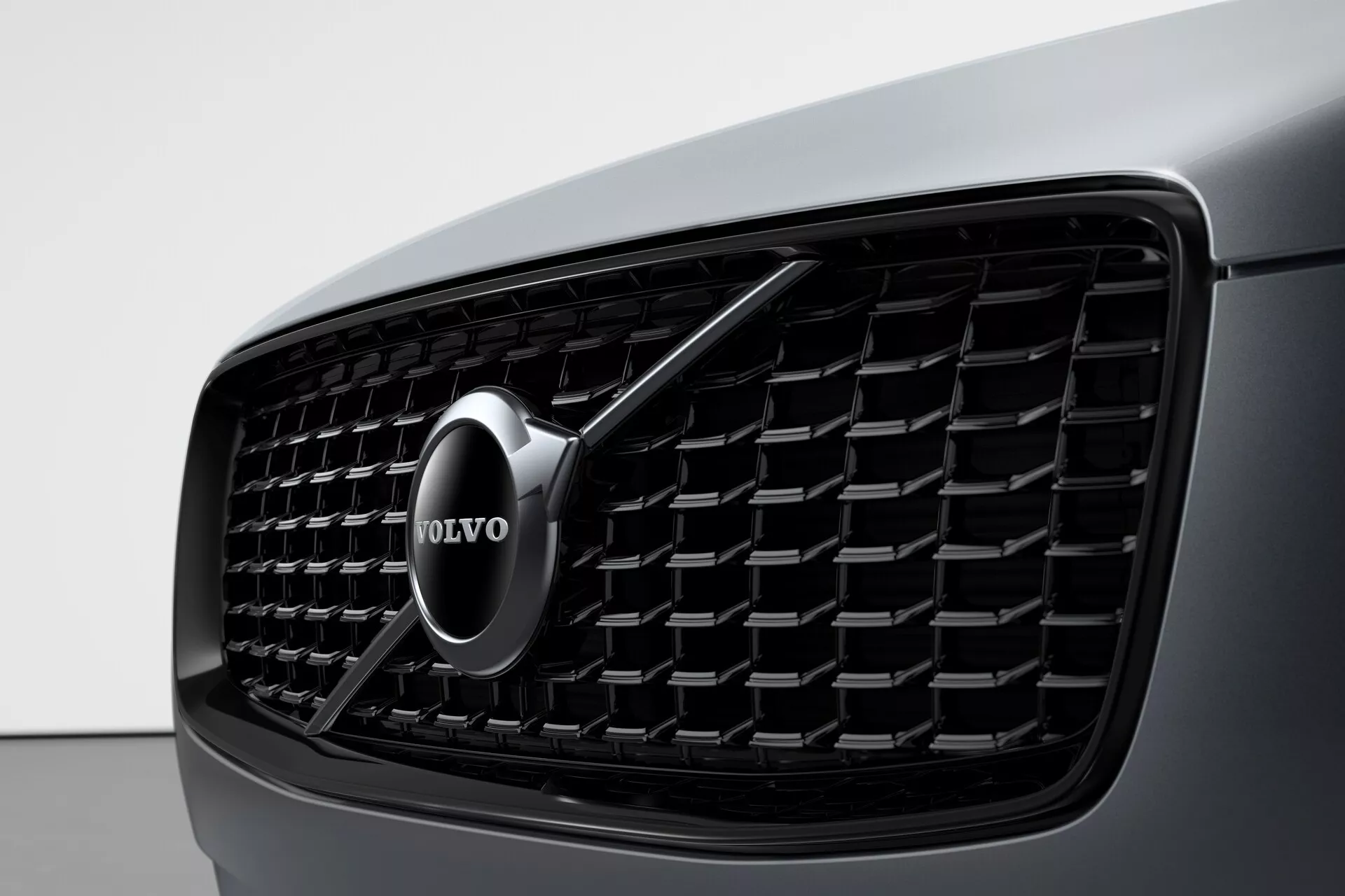 Primo piano della sezione frontale di una Volvo XC90 2015 (2ª generazione) che si concentra sulla griglia, caratterizzata da un fitto motivo a rete con elementi rettangolari, un emblema Volvo cromato al centro con una freccia che punta diagonalmente verso l'alto a destra e una vista della carrozzeria scura e slanciata dell'auto a sinistra. L'immagine mette in risalto l'estetica del design e il riconoscimento del marchio.