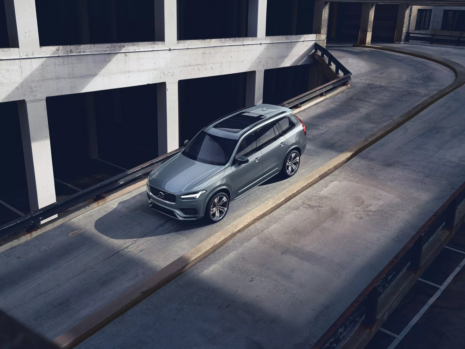 Vista dall'alto angolare di un SUV Volvo XC90 del 2015 parcheggiato su una superficie di cemento, che mostra il suo design elegante e moderno con finitura in vernice metallizzata, caratteristica griglia anteriore, fari visibili e cerchi in lega intricati. Lo scatto mette in risalto il tetto, il cofano e il lato sinistro dell'auto, con l'ombra proiettata che suggerisce luce del mattino o del tardo pomeriggio. L'ambiente sembra essere un livello superiore di una struttura di parcheggio a più piani, esaltando la prominenza del veicolo in un ambiente urbano pulito.